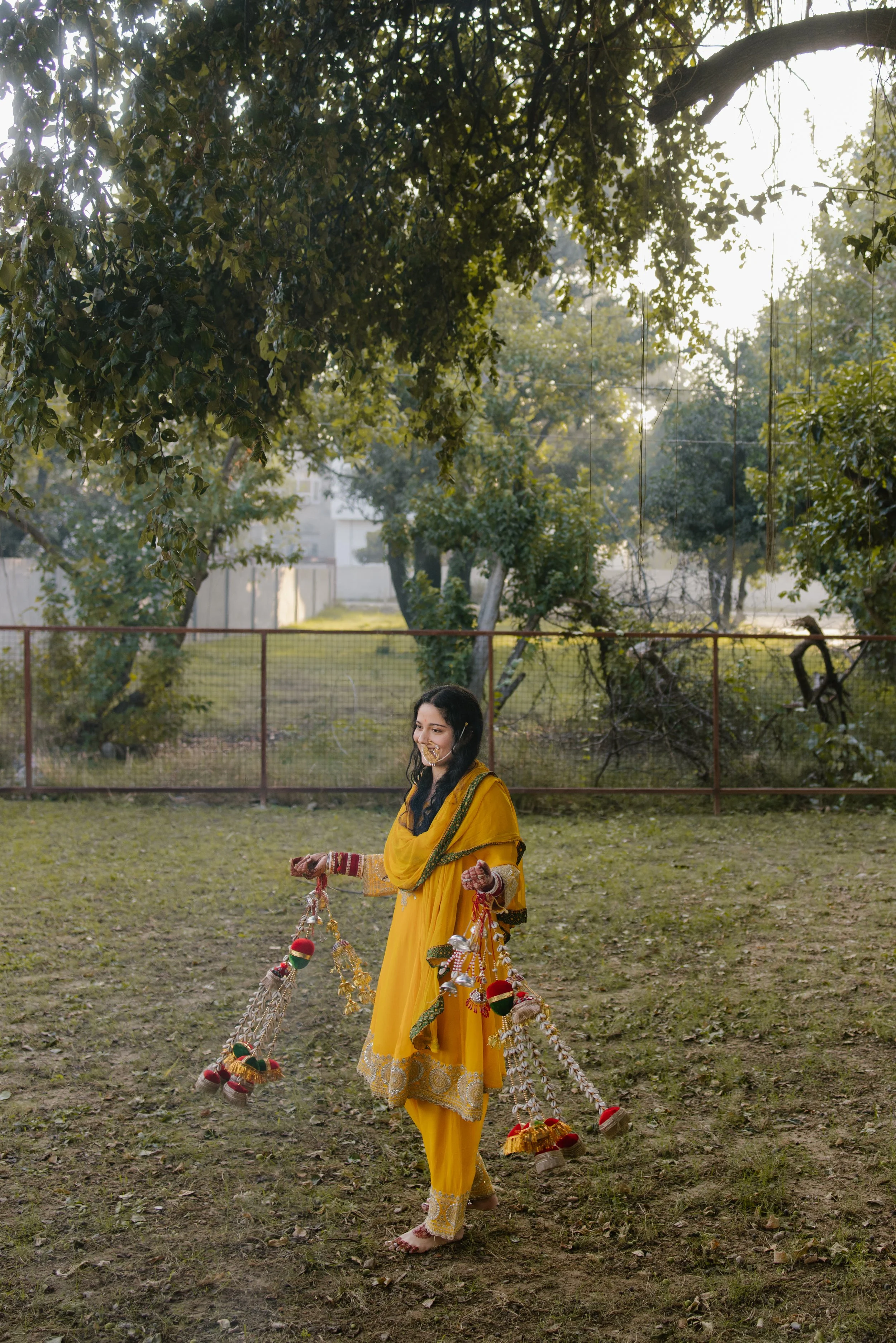 A woman in traditional yellow Indian attire with gold accents, holding colorful decorative items, standing outdoors in a grassy area with trees and a fence in the background, during daylight.