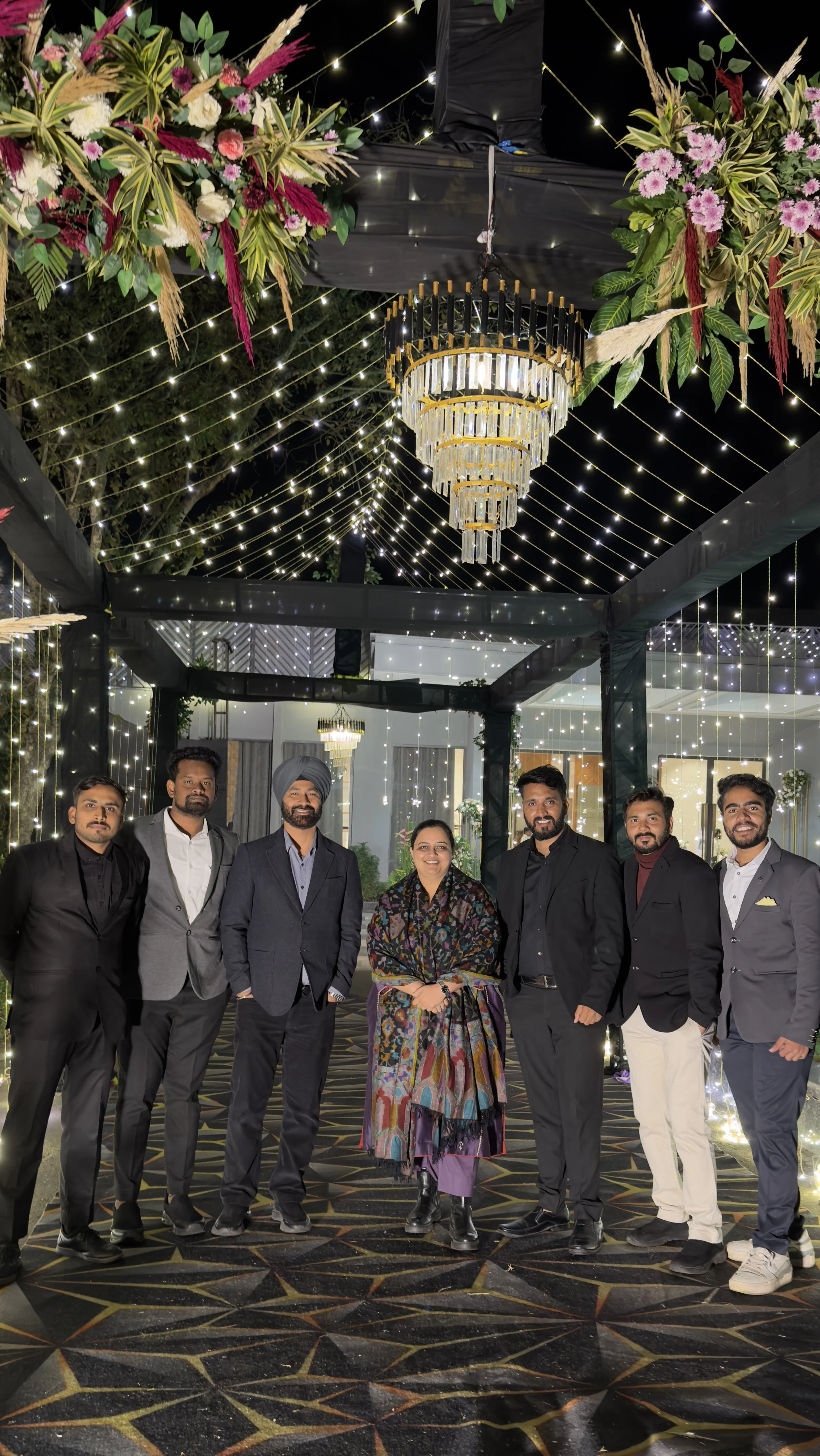 A group of seven people, six men and one woman, standing together at an event with a decorated ceiling featuring string lights and floral arrangements, posing for the photo.