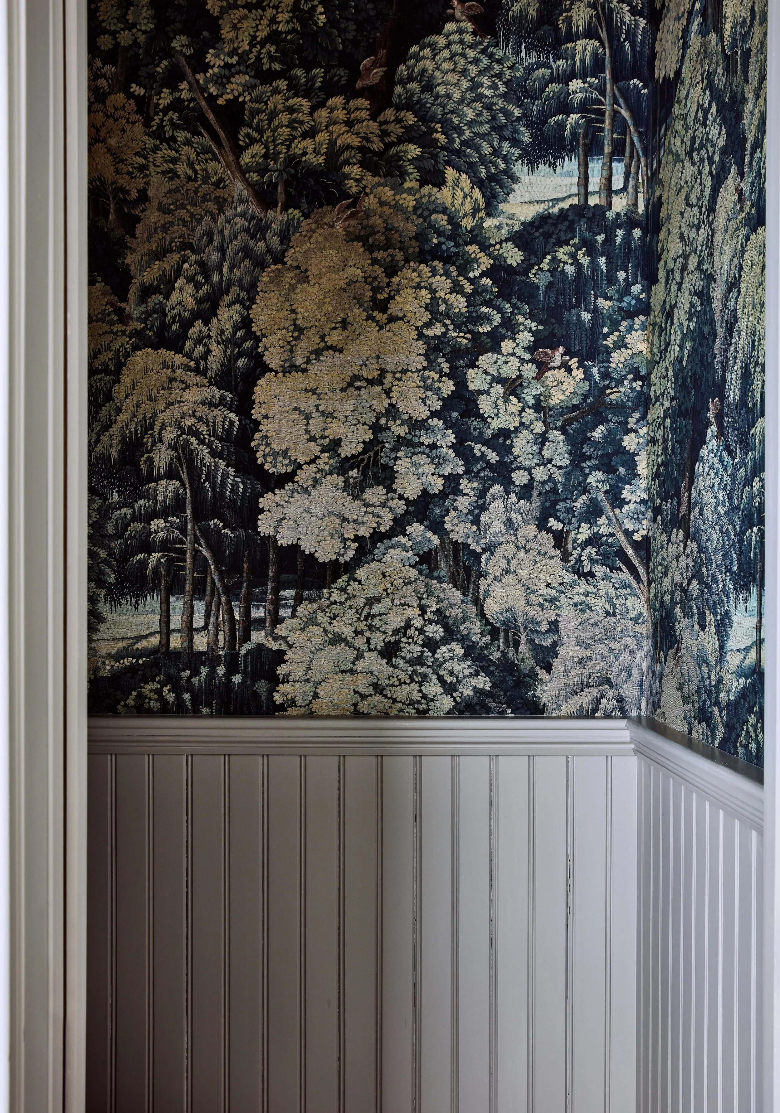 Interior wall with a traditional tapestry or wallpaper depicting a dense forest scene with trees and bushes in shades of green and beige, combined with white beadboard wainscoting at the bottom.