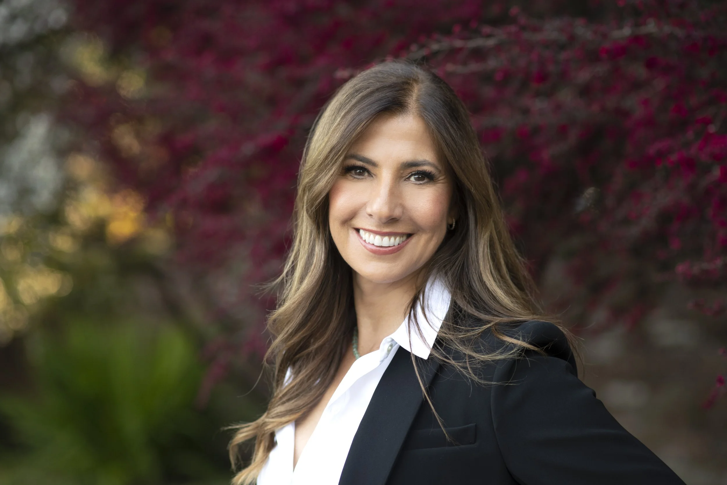 A woman with long brown hair smiling, wearing a black blazer and white shirt, standing outdoors with trees and red leaves in the background.