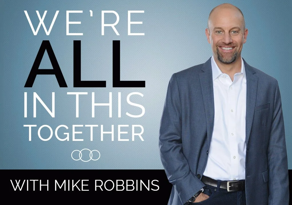 A smiling man in a suit standing in front of a blue background with text "WE'RE ALL IN THIS TOGETHER" and "WITH MIKE ROBBINS."
