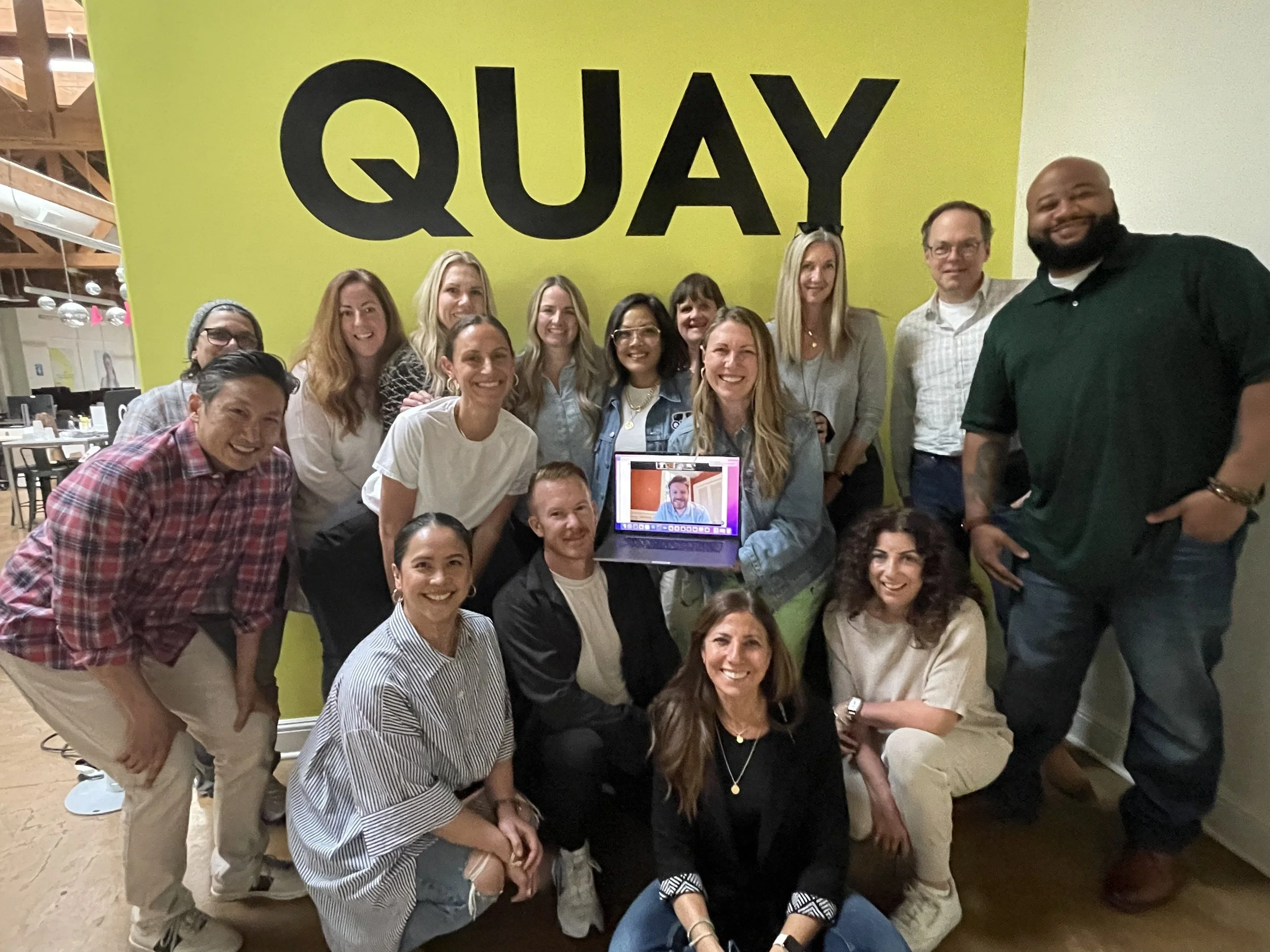 Group of 16 people gathered in front of a large yellow wall with the word 'QUAY' written on it. They are smiling and posing for a photo indoors, with some kneeling in front and others standing behind. A laptop with a video call is visible among them.