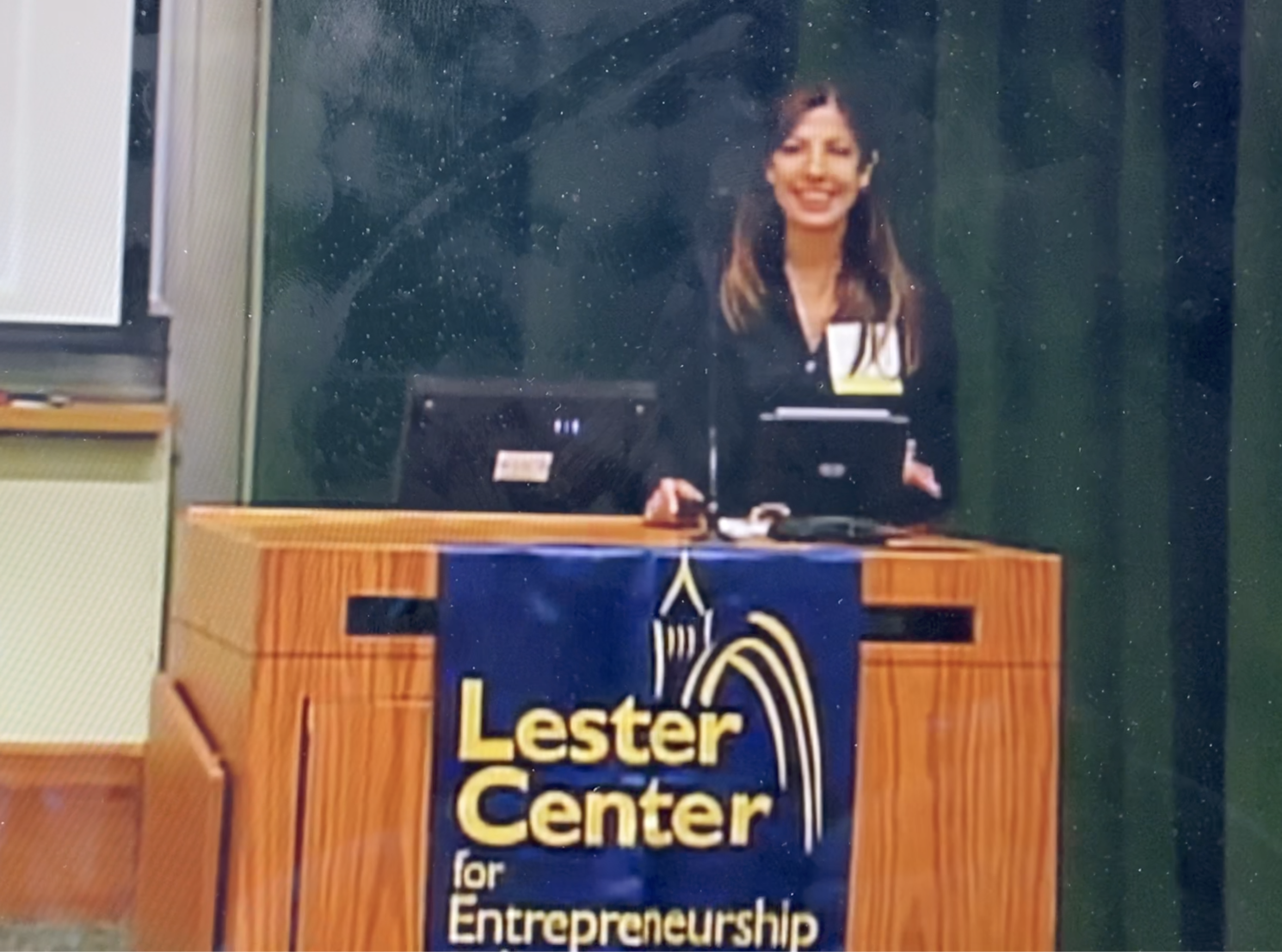 A woman standing behind a wooden podium with a blue and yellow sign that reads "Lester Center for Entrepreneurship." She is smiling and wearing a black jacket in a classroom with a chalkboard behind her.