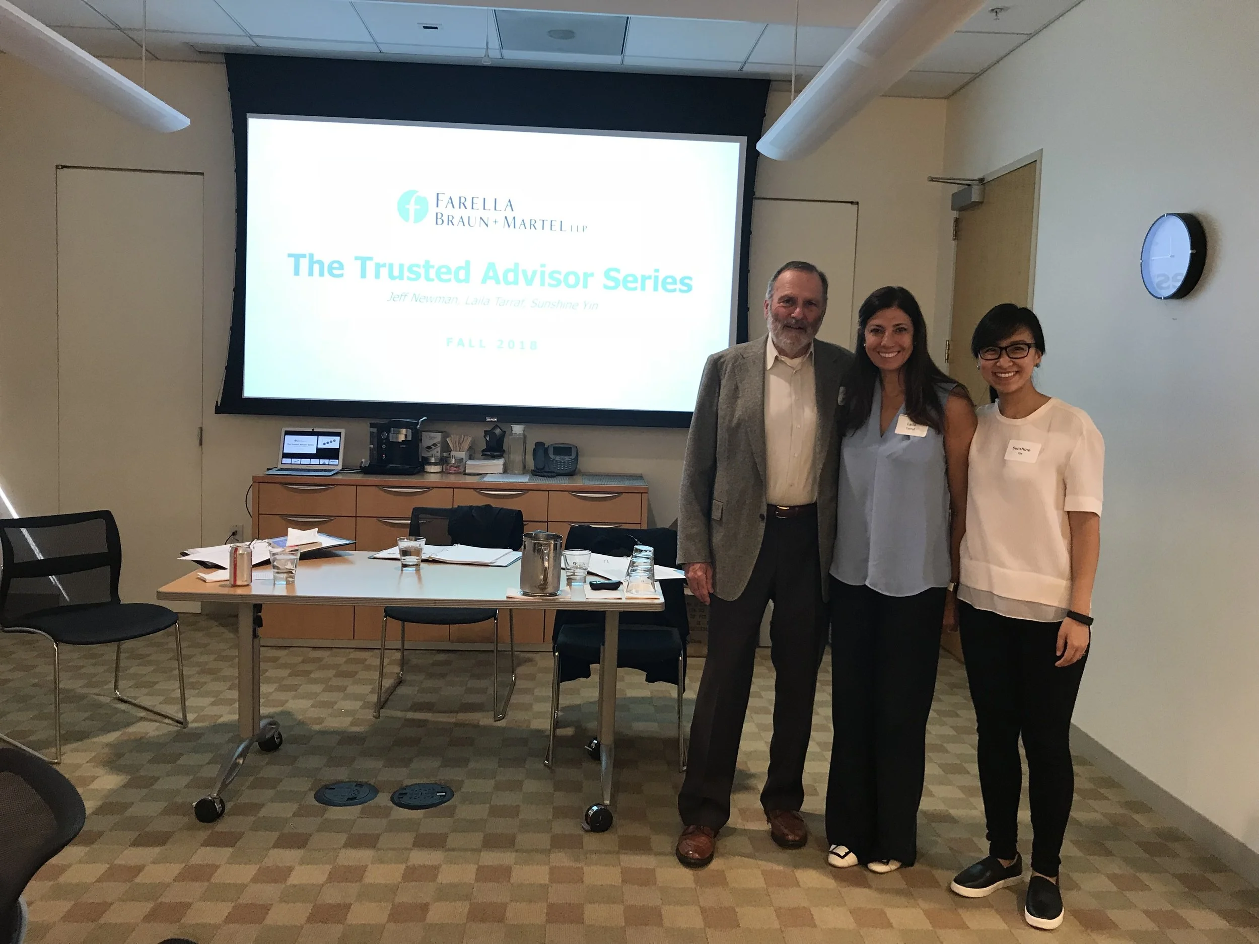 Three people standing in front of a presentation screen in a conference room. The screen displays a logo and text indicating a session called 'The Trusted Advisor Series' with fall 2018 date. Conference table and chairs are visible, along with laptops, glasses, and snacks on the table.