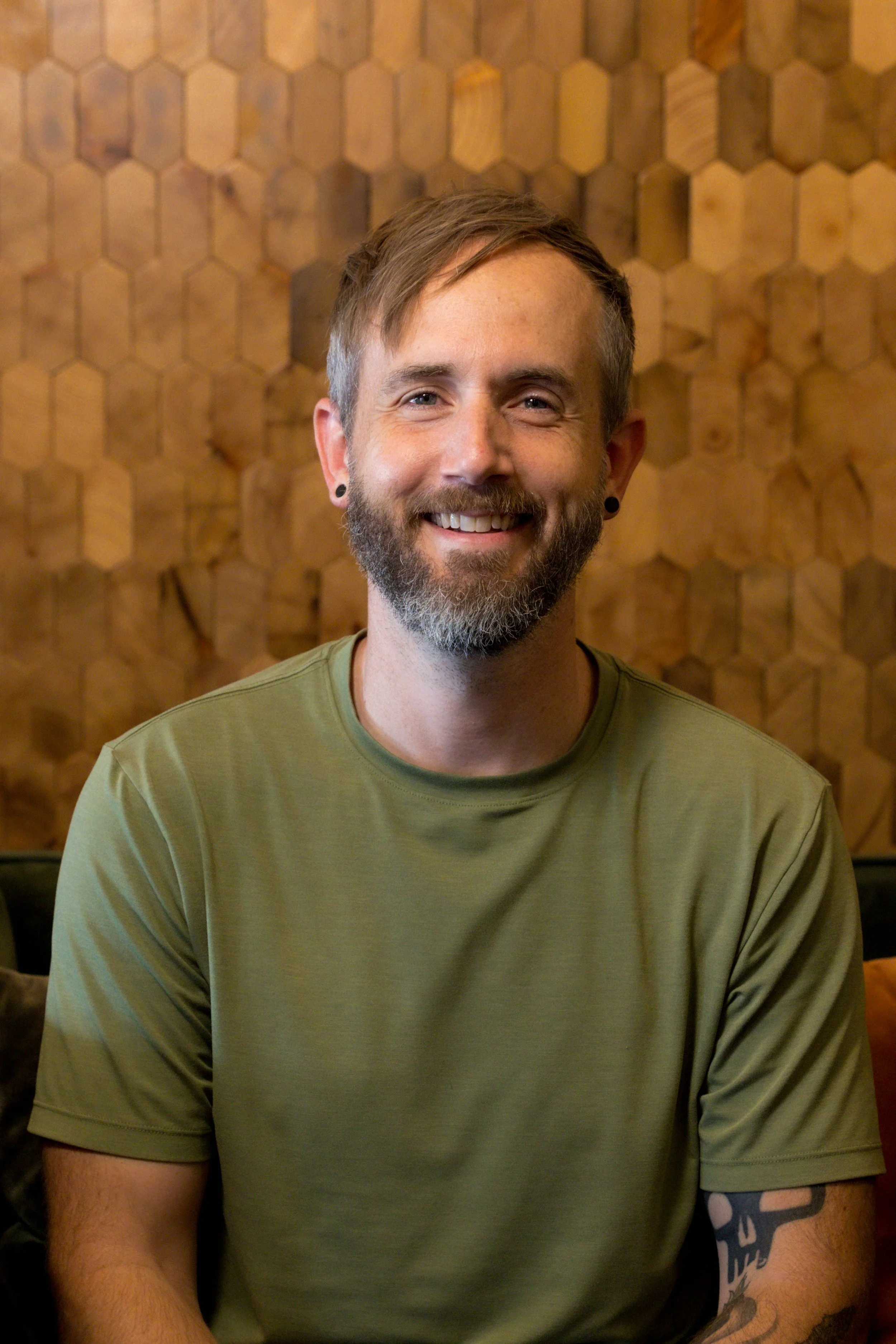 A smiling man with a beard, wearing an olive-green T-shirt, sitting in front of a wooden honeycomb-patterned wall. He is Nic, a massage therapist at Very Well KC.