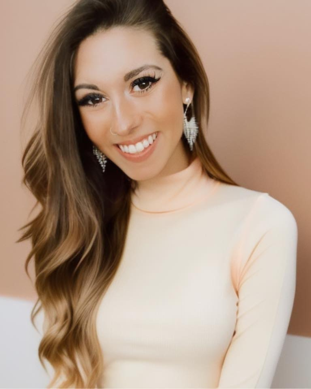 A smiling woman with long, wavy brown hair wearing a light-colored top, large dangling earrings, and a nose ring, against a plain background.