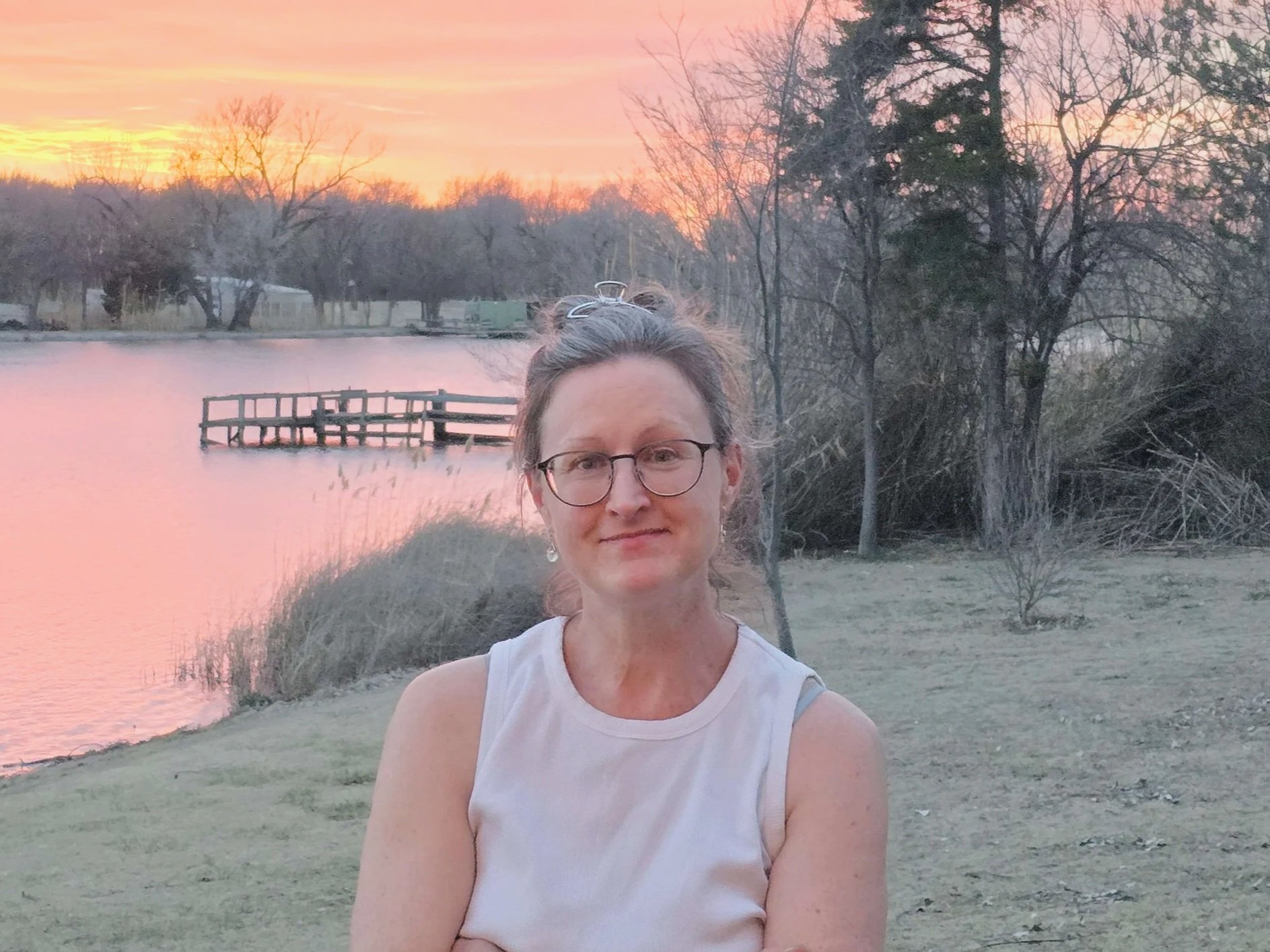 A woman with glasses and earrings sitting outdoors during sunset near a lake with trees and a small dock in the background.