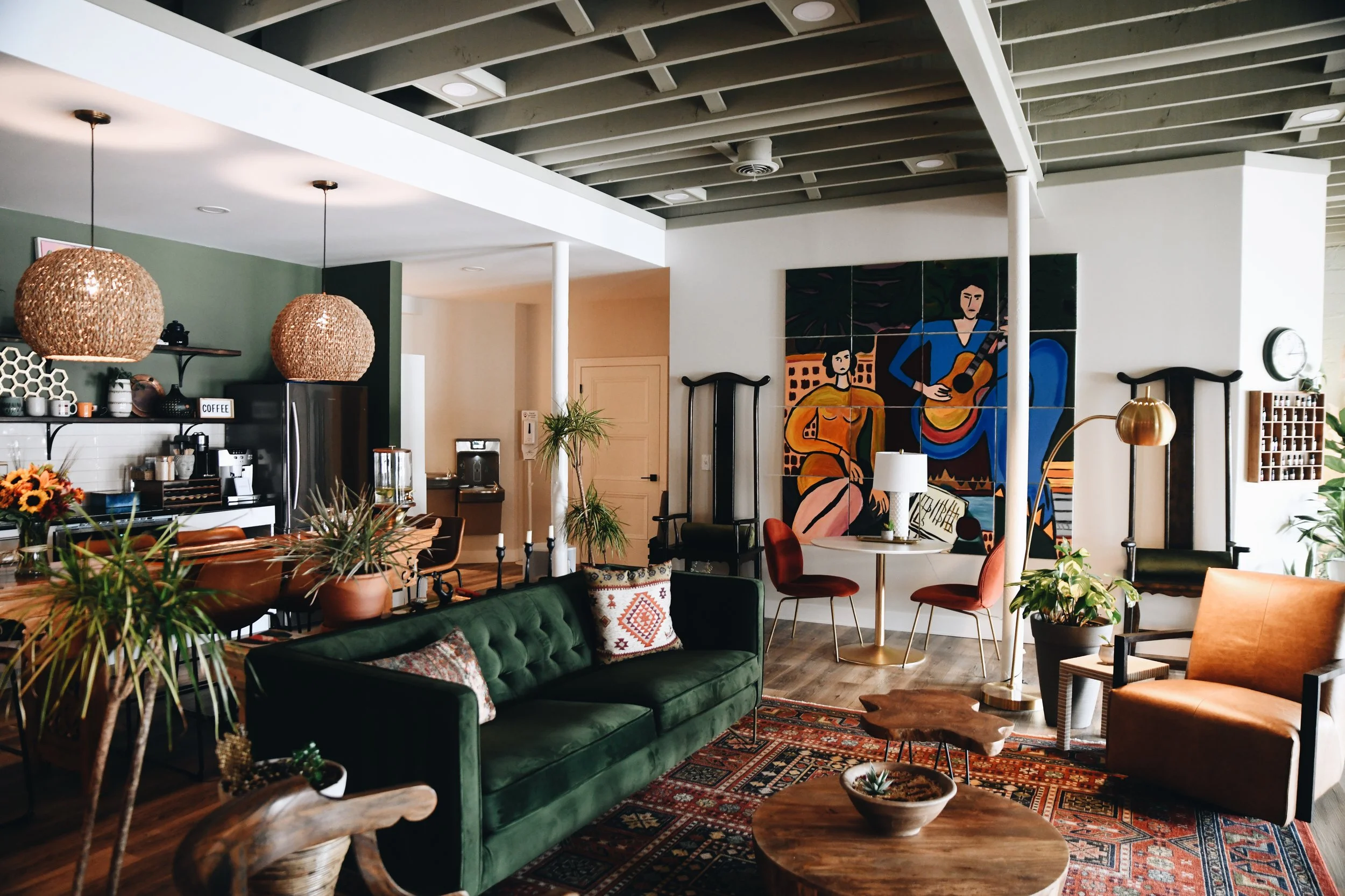 A cozy living room with a green velvet sofa, wooden coffee tables, colorful cushions, and a vibrant abstract painting on the wall. There are various plants and modern lighting fixtures, including hanging wicker lamps, creating a warm atmosphere.