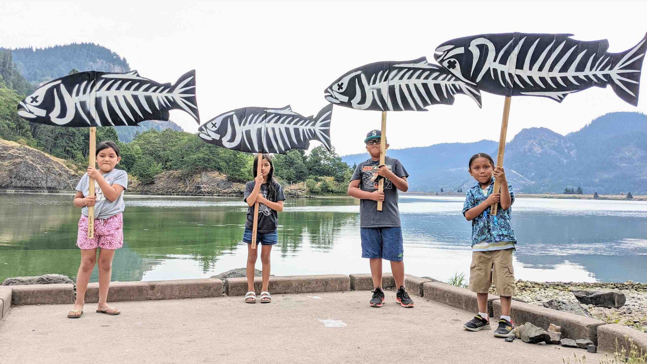 Standing Vigil on the Little White Salmon River. Alana Harvey age 6, Mila Gandara age 8 , Tyler Takala age 10, and Clint Takala age 6 hold cardboard cutout signs painted to look like salmon. 