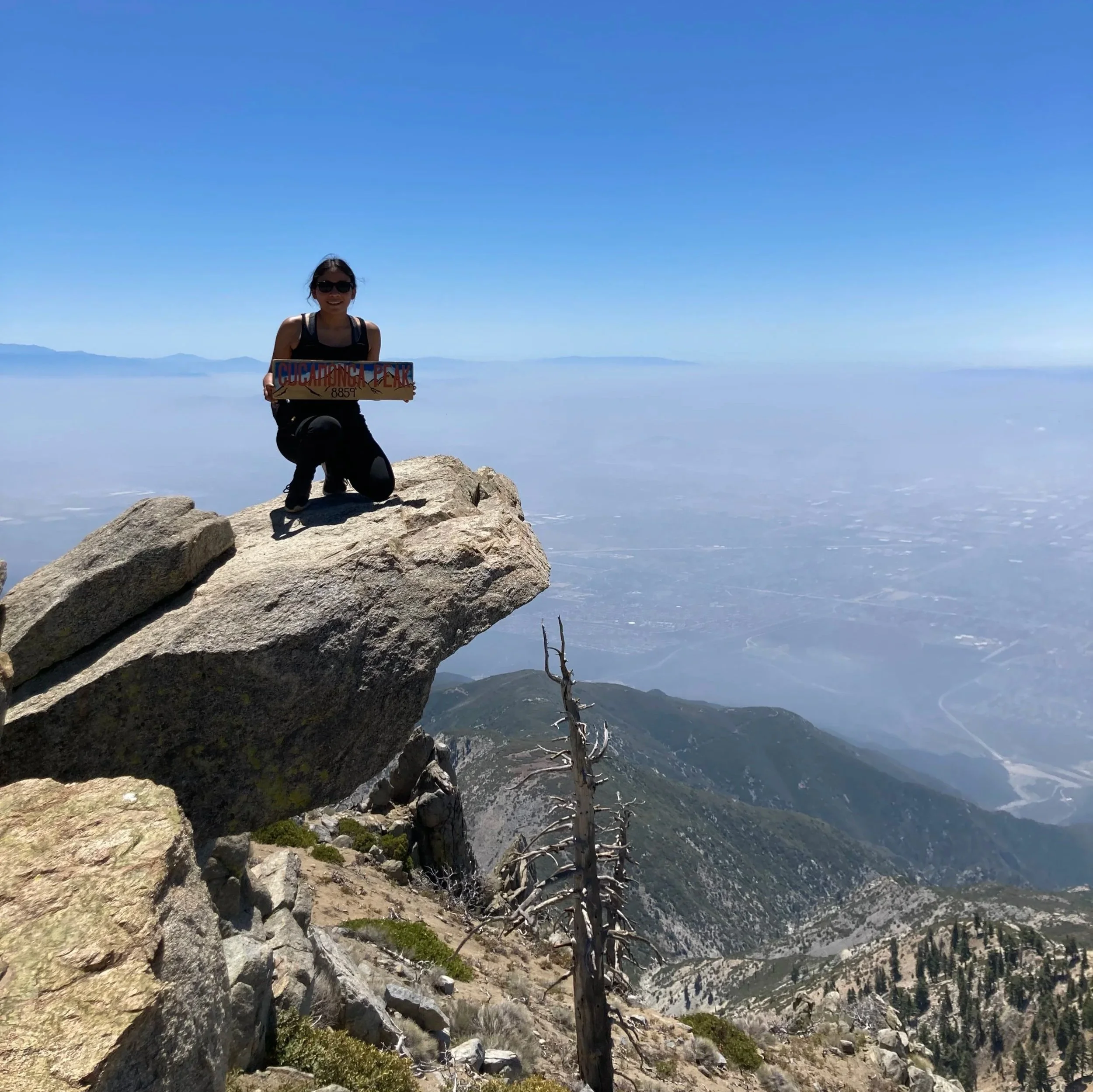 A woman in sunglasses kneeling on a large rock at the summit of a mountain, holding a sign that reads 'Cucamonga Peak 8859 ft'. The background shows a clear blue sky, distant mountains, and a valley below.
