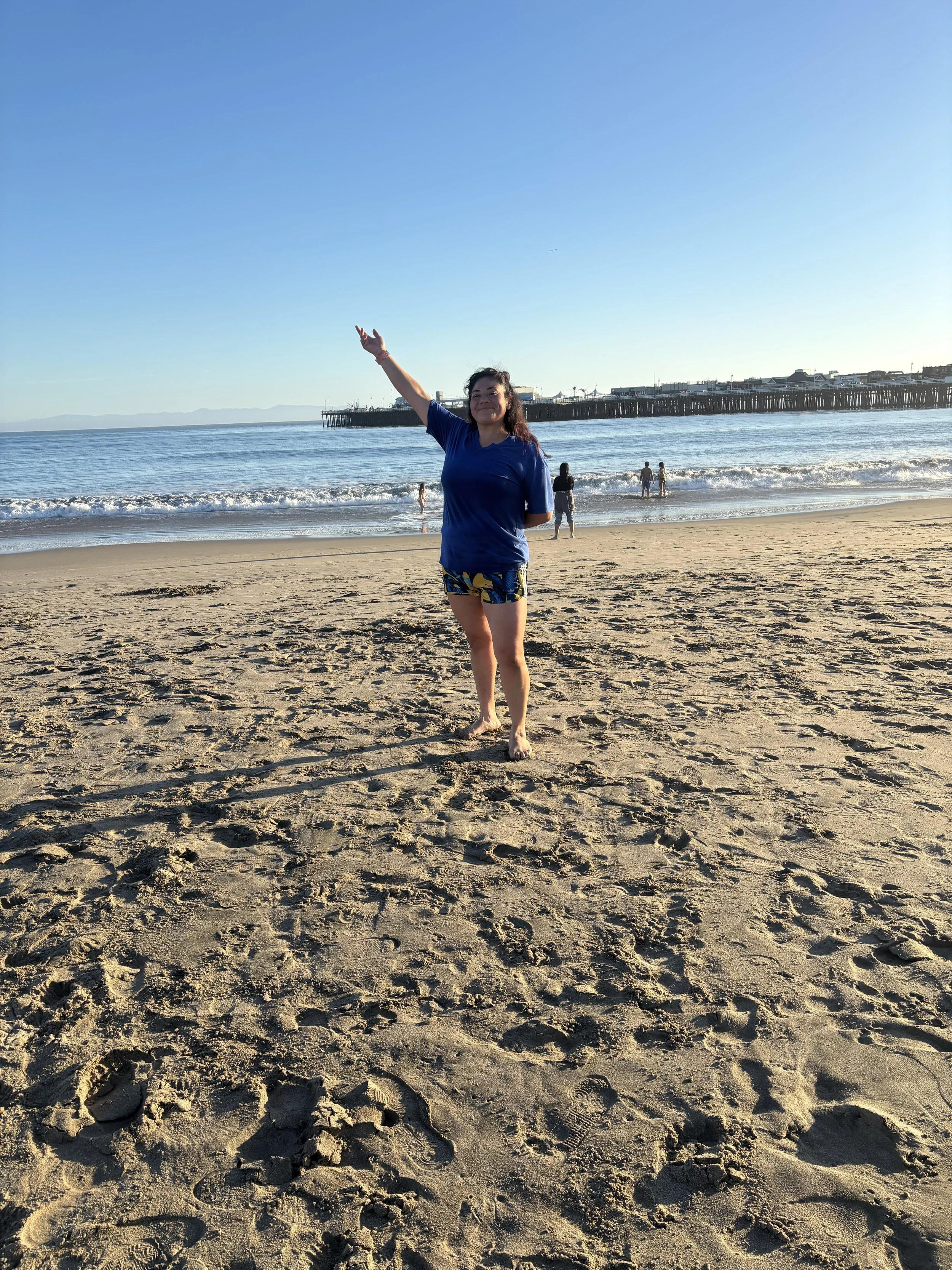 A woman standing on a sandy beach with her arm raised, smiling, wearing a blue shirt and patterned shorts. In the background, there are people in the ocean, a pier extending into the water, and a clear sky.