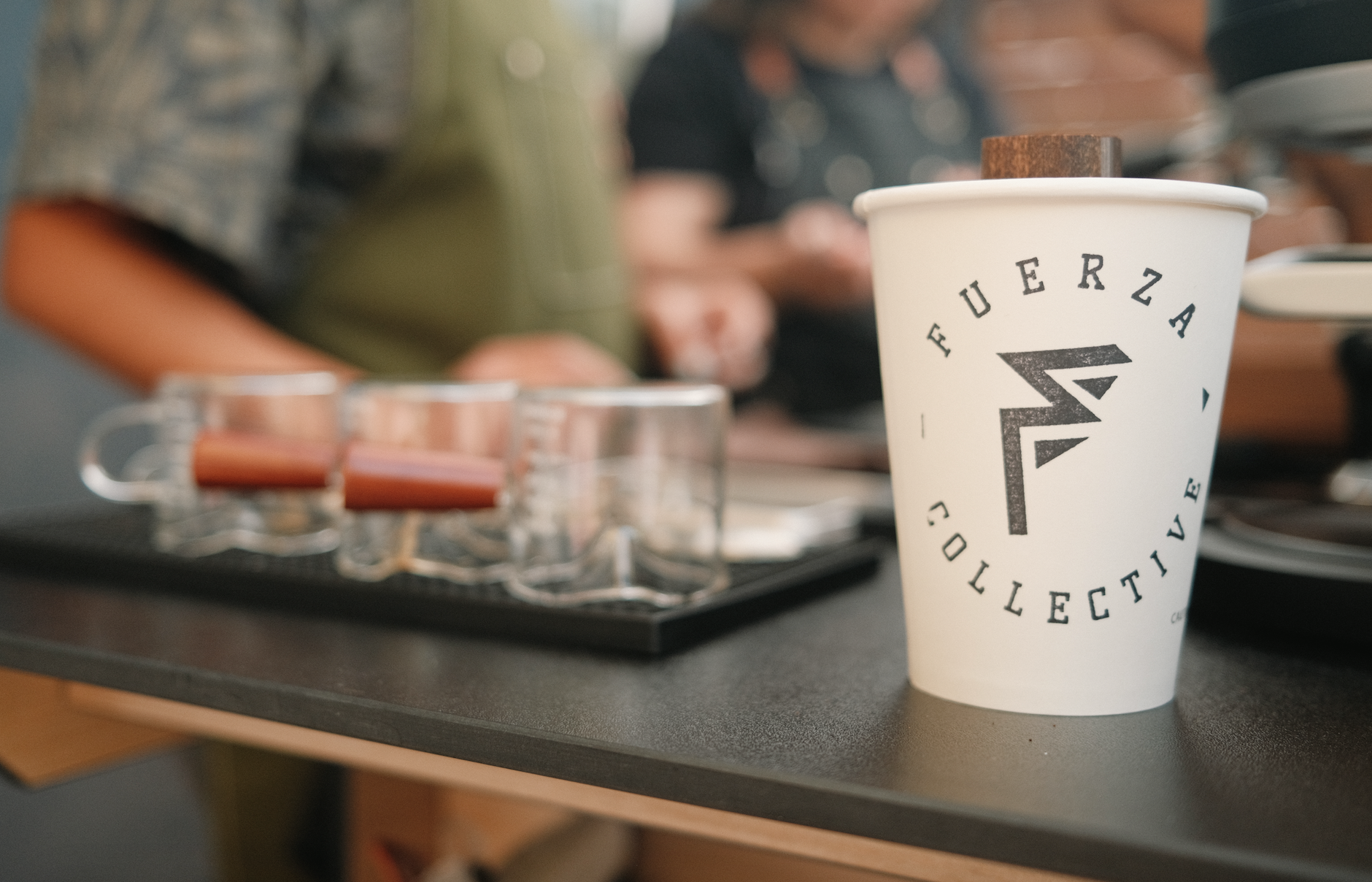 A disposable coffee cup with the FUERZA collection logo on a countertop, with blurred people in the background and a tray of shot glasses with orange liquid.
