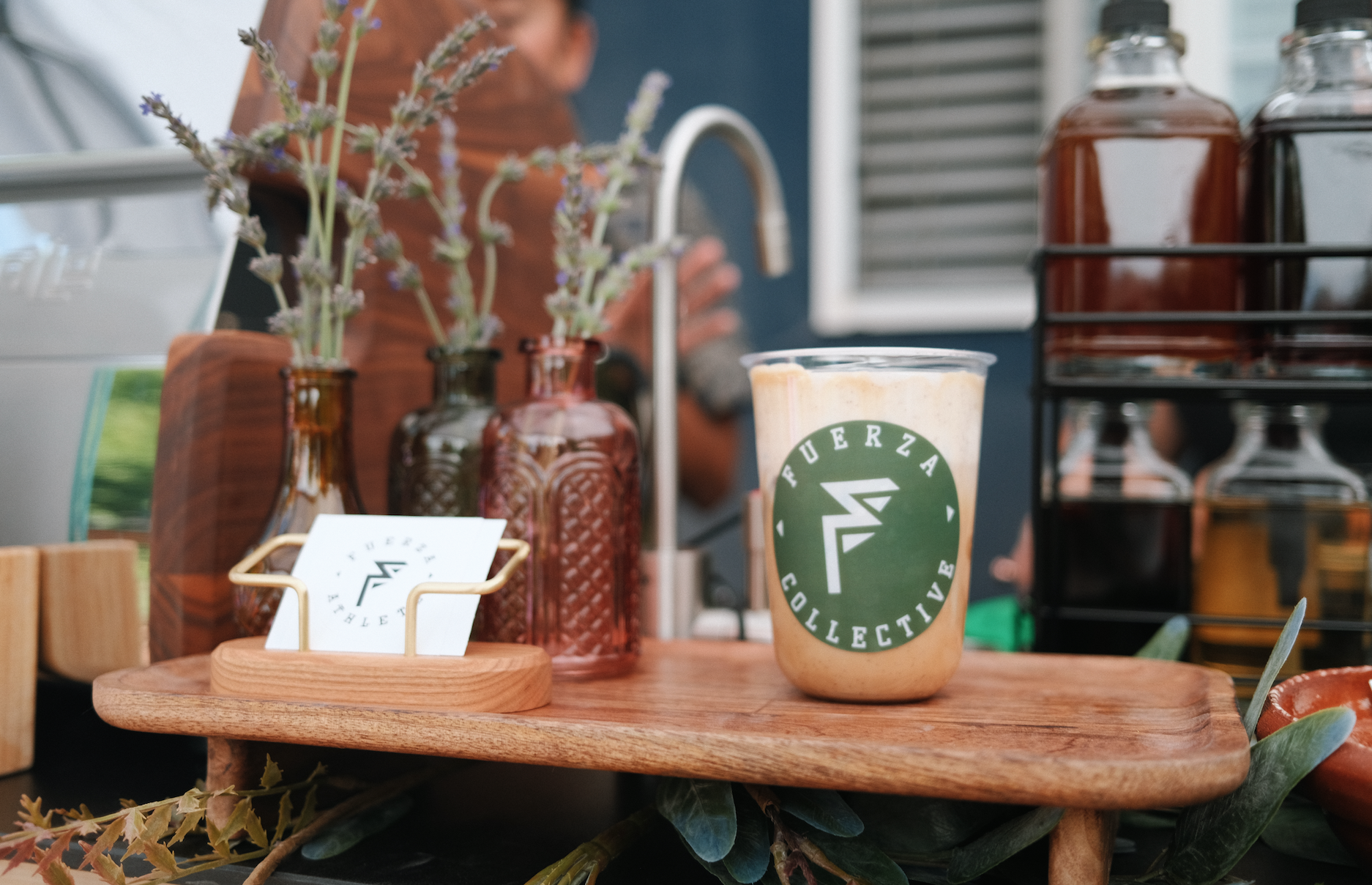 A wooden serving tray with a pint of coffee or smoothie with a green logo that reads 'Fuerza Collective,' and decor including amber and clear glass bottles with lavender and other small flowers, placed on a counter with a person in the background near a sink and window.