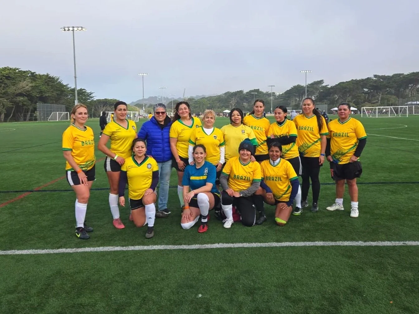 Women football players in yellow jerseys with green accents, and a few in blue jerseys, posing on a soccer field with a coach, during a game. The field has goal posts, surrounded by trees and tall light poles.