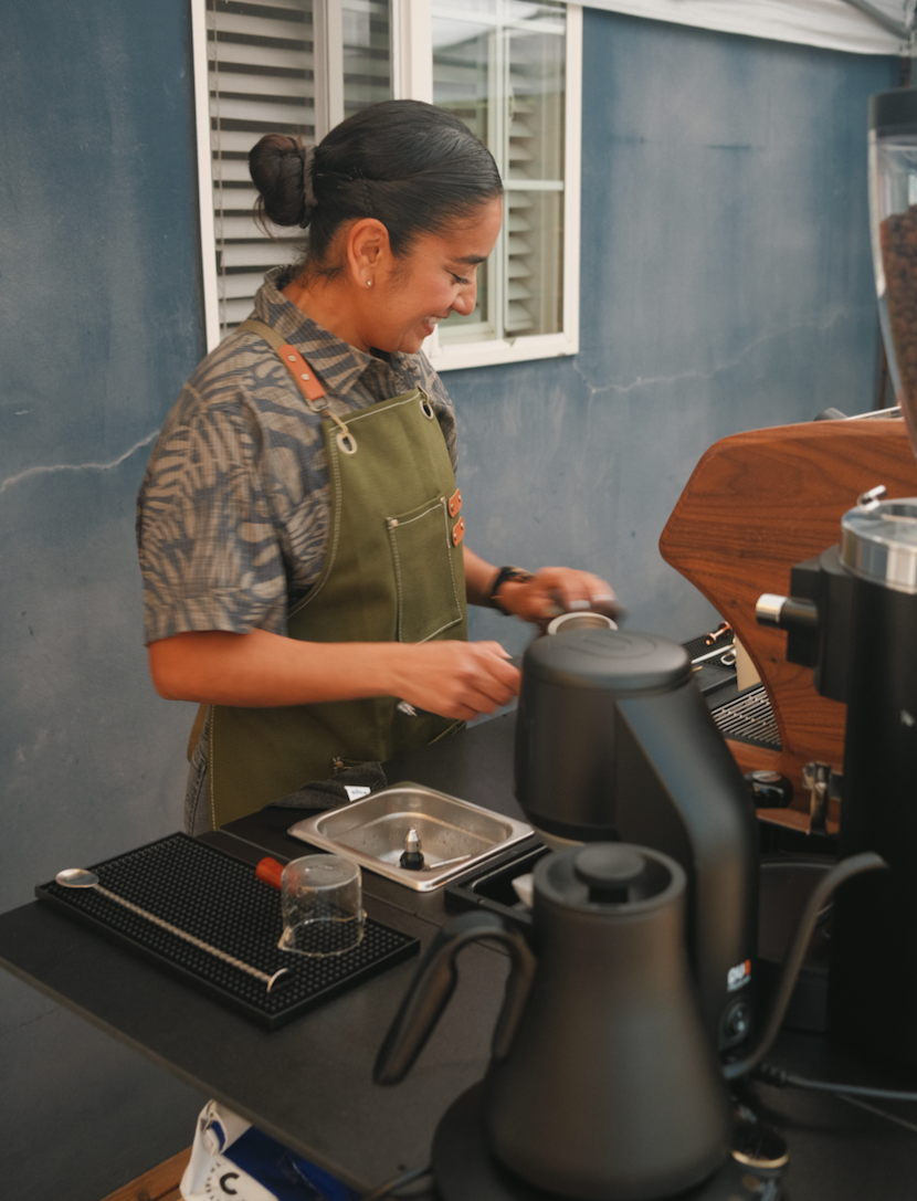 Owner and barista Christina, smiling, wearing a patterned shirt and an apron, working with a La Spaziale espresso machine and brewing accessories on a black top mobile coffee cart.