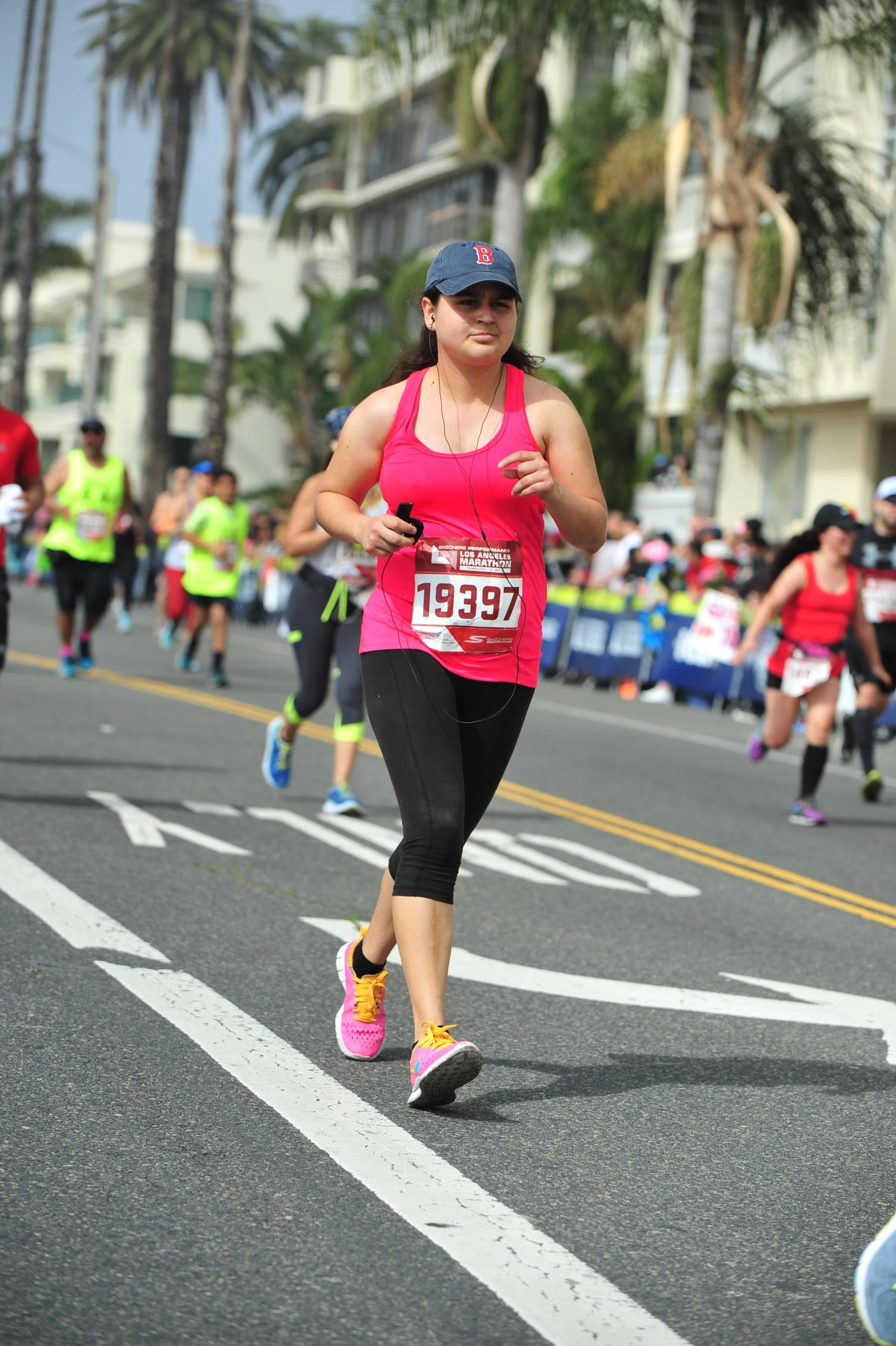 A woman running in a marathon wearing a pink top, black leggings, pink and yellow sneakers, a dark cap, and headphones, with other runners and palm trees in the background.