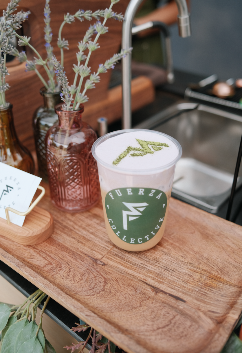 A Fuerza Collective iced coffee with cold foam and matcha on top, in a 8oz cup with a Fuerza Collective logo, placed on a wooden surface with decorative lavender in glass bottles nearby.
