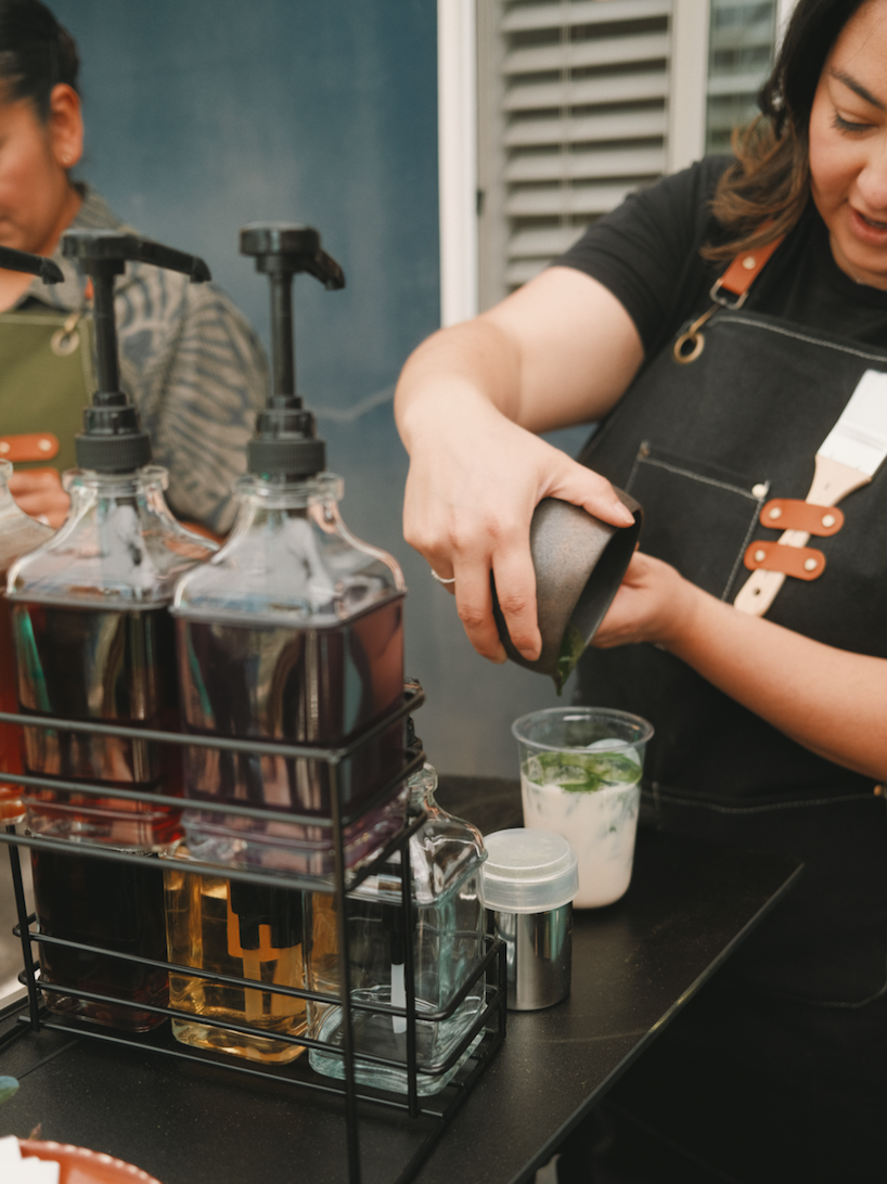 A Fuerza Collective barista pouring premium matcha into a glass of iced drink on a mobile coffee cart, with bottles of syrups or flavorings on the side.