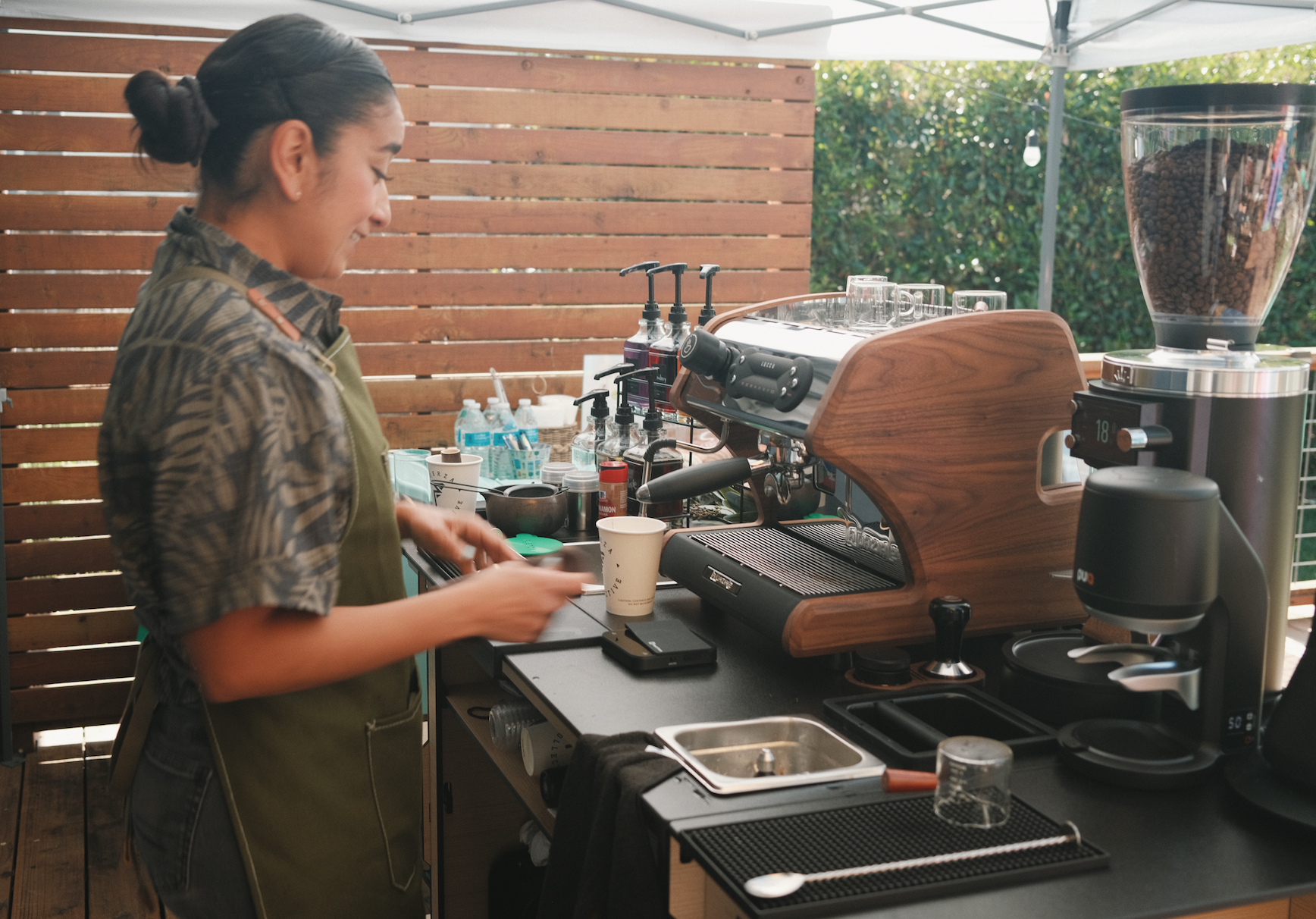 Owner and barista Christina preparing coffee behind an espresso machine on a mobile coffee cart, with a wooden privacy fence and greenery in the background.