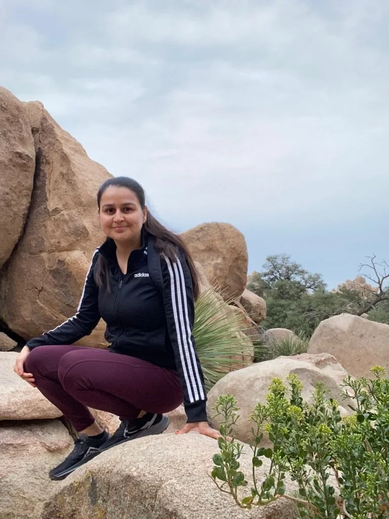 A woman in a black Adidas jacket and maroon pants crouches on a large rock in a desert landscape with scattered shrubs and large boulders, under a cloudy sky.
