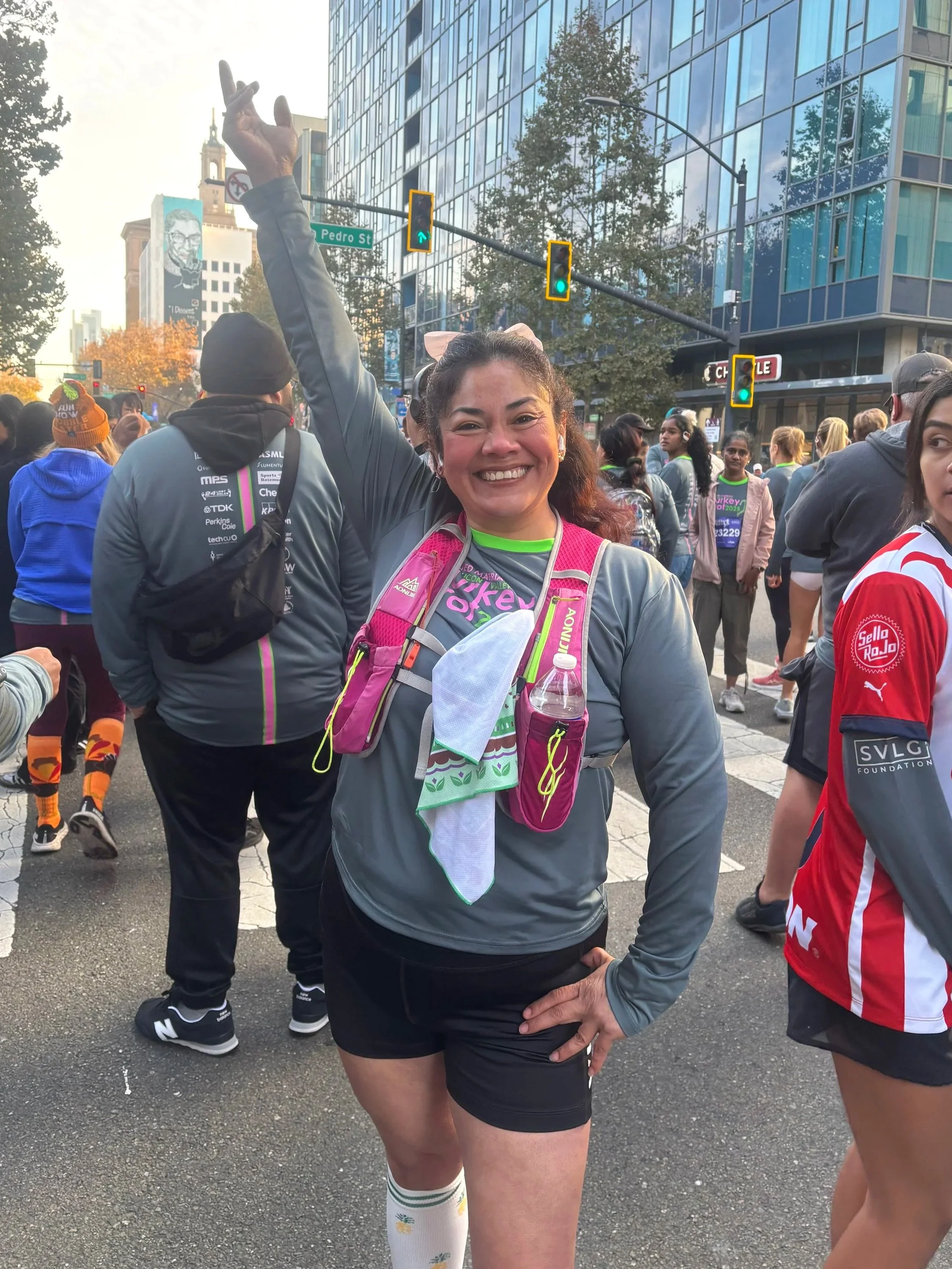Smiling woman in athletic clothing standing in a crowded city street during a race, with runners and tall buildings in the background.