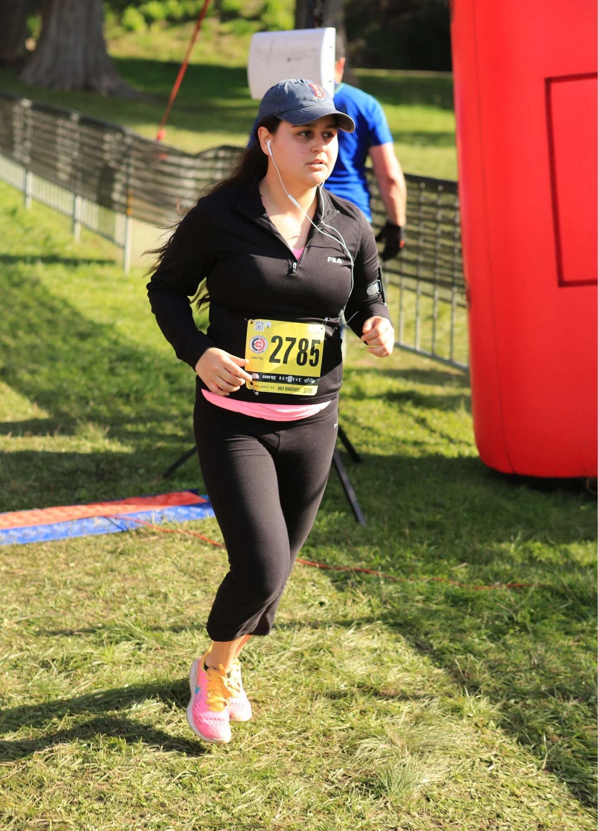 A young woman running outdoors in a marathon, wearing a black jacket, black leggings, pink running shoes, and a baseball cap, with a race bib numbered 2785.