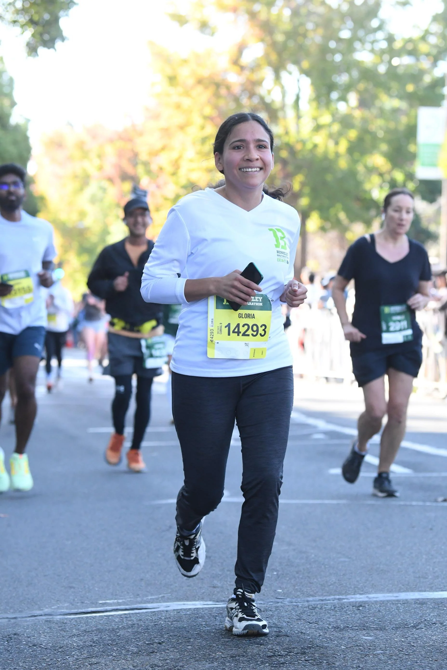 A woman running in a marathon, smiling, wearing a white long-sleeve shirt, black pants, and sneakers, holding a phone, with other runners behind her and trees in the background.