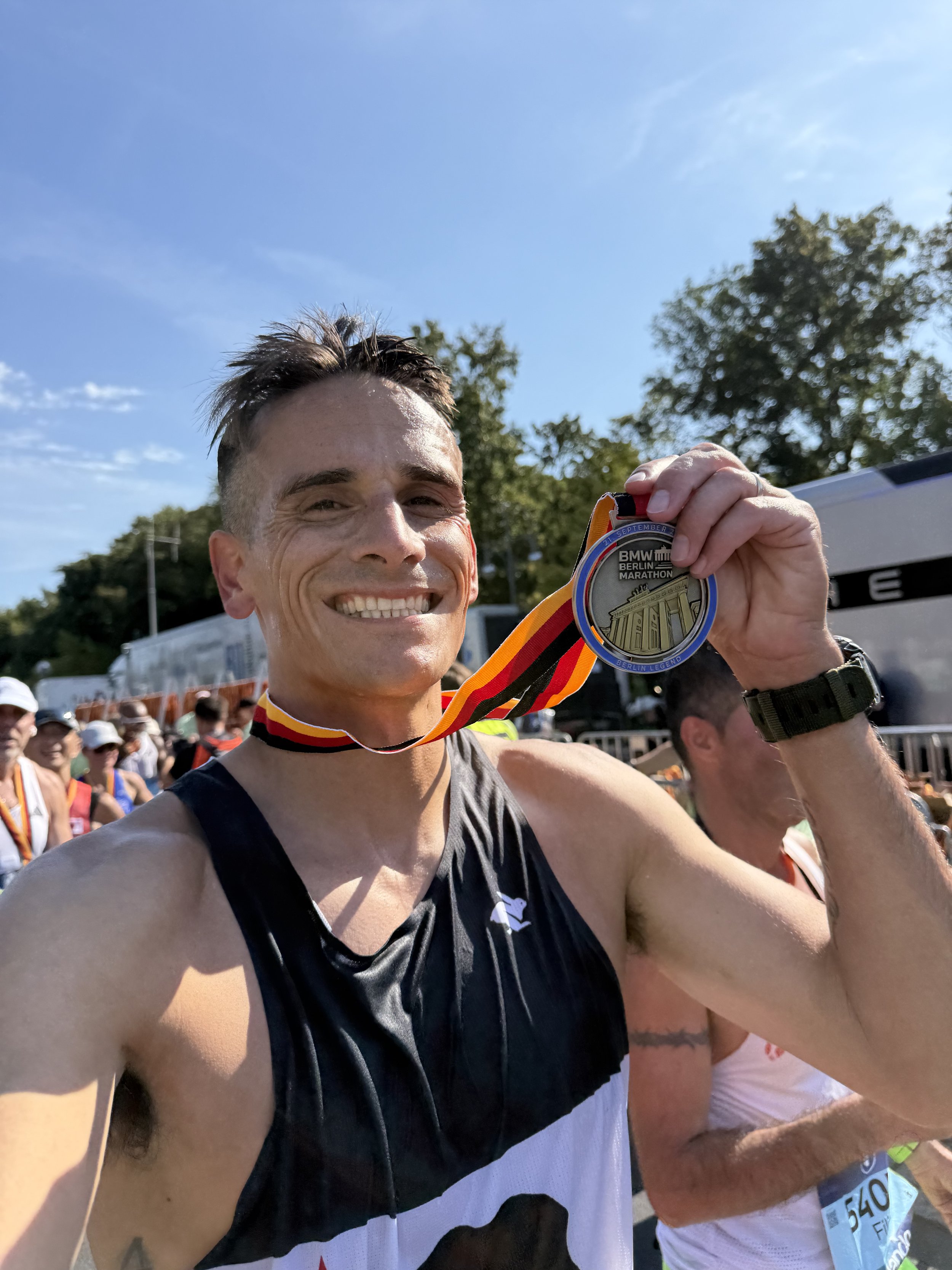 Smiling male marathon runner showing a finisher's medal at the Berlin Marathon, with other runners and trees in the background on a sunny day.