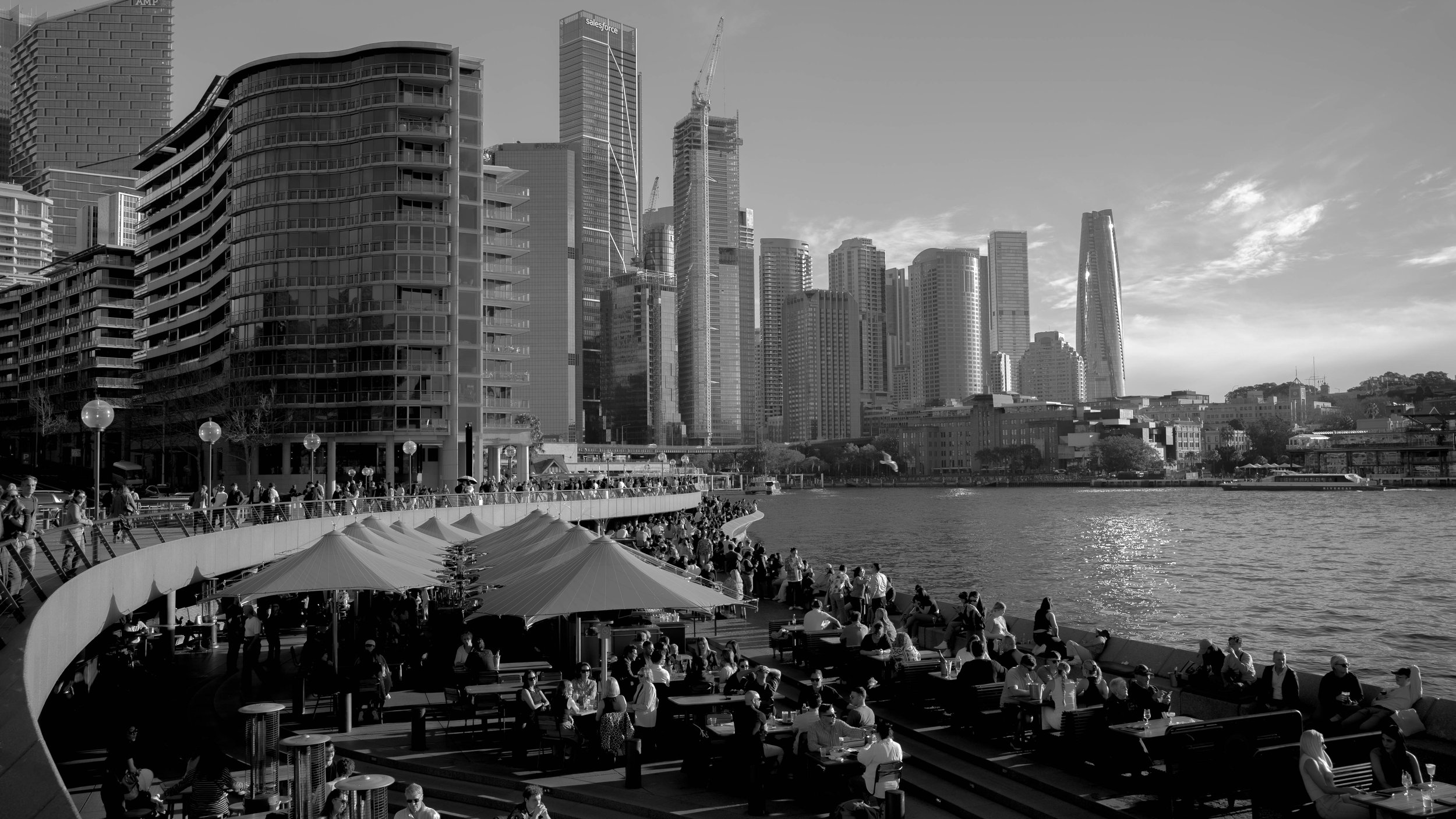 A black and white photo of Sydney, Australia's waterfront with modern skyscrapers in the background, a promenade with people walking, and an outdoor dining area with tables, umbrellas, and people sitting.