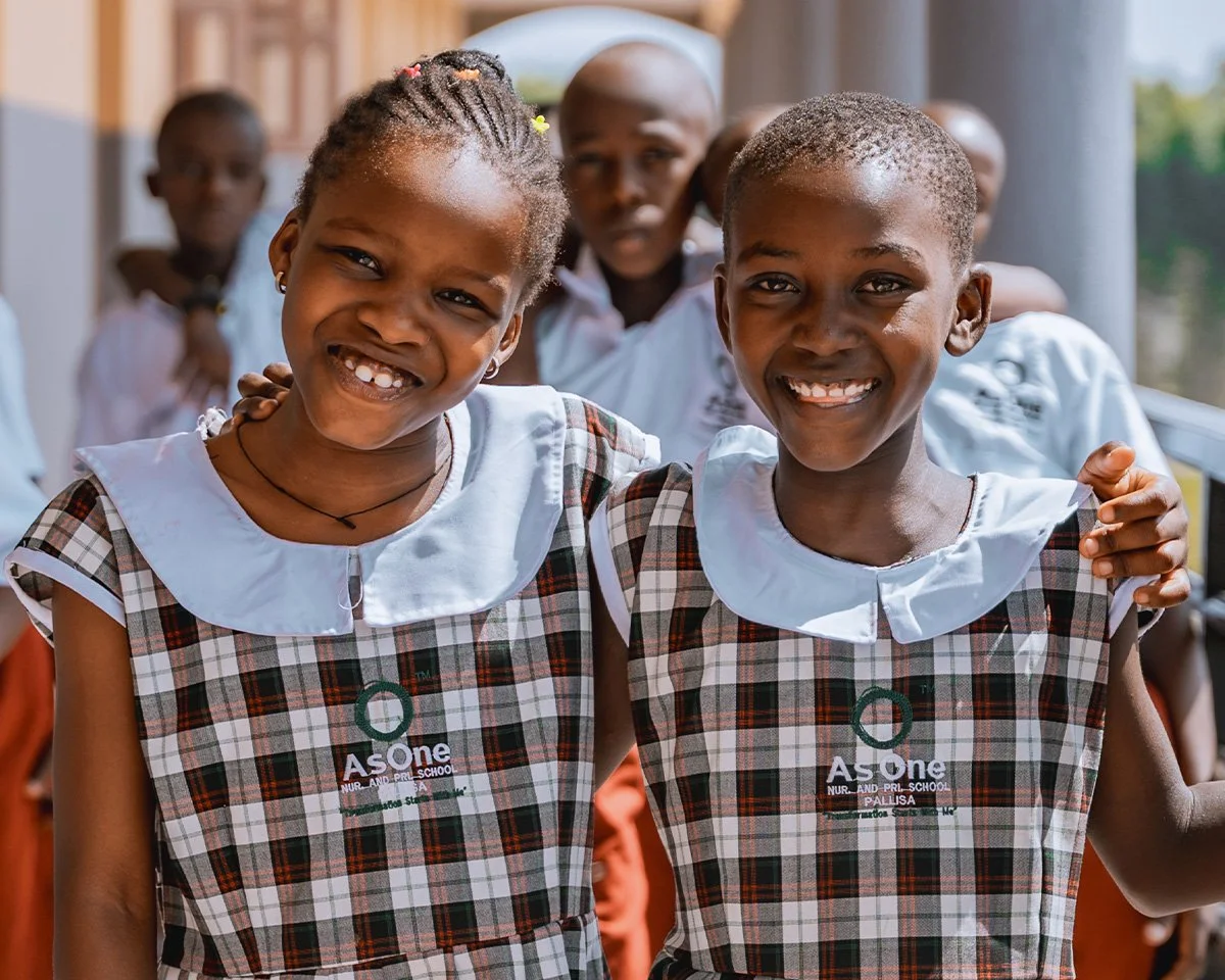 Two smiling AsOne Nursery & Primary School, Pallisa students in uniform standing arm in arm