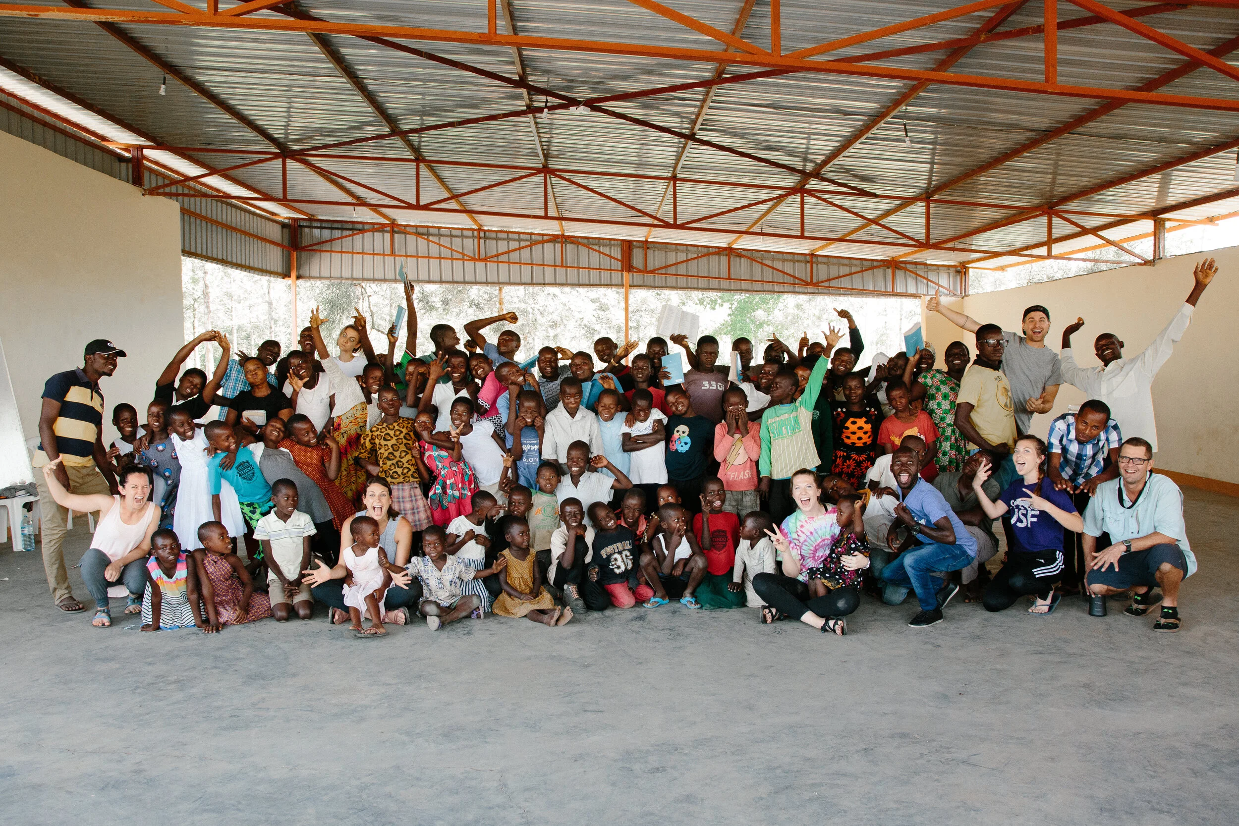 Large group photo of AsOne immersion trip participants with a crowd of Ugandan children, celebrating together under a covered pavilion