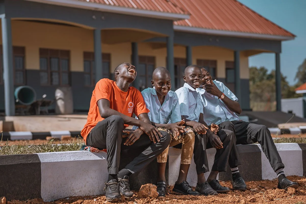 Four AsOne High School students in Kameramaido laughing together on a painted curb outside their school building, one in an orange AsOne shirt and three in light blue uniforms