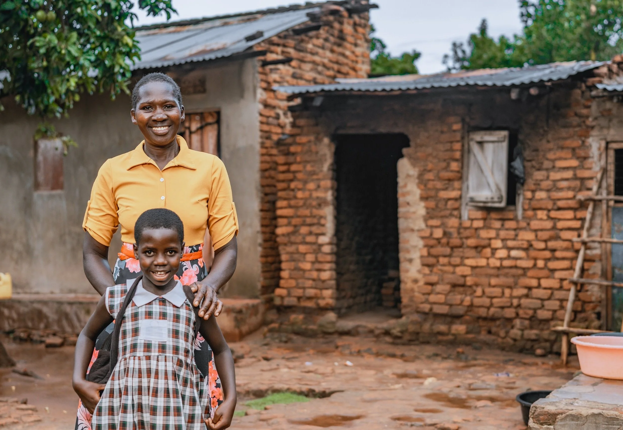 AsOne parent standing with her daughter in school uniform outside their home in Idudi, Uganda
