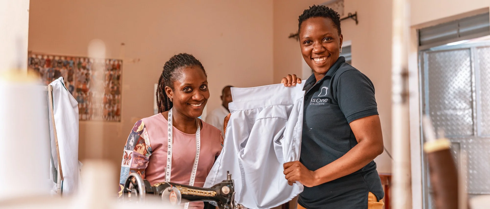 AsOne staff member and a tailor smiling while holding a finished shirt at the tailoring center in Idudi