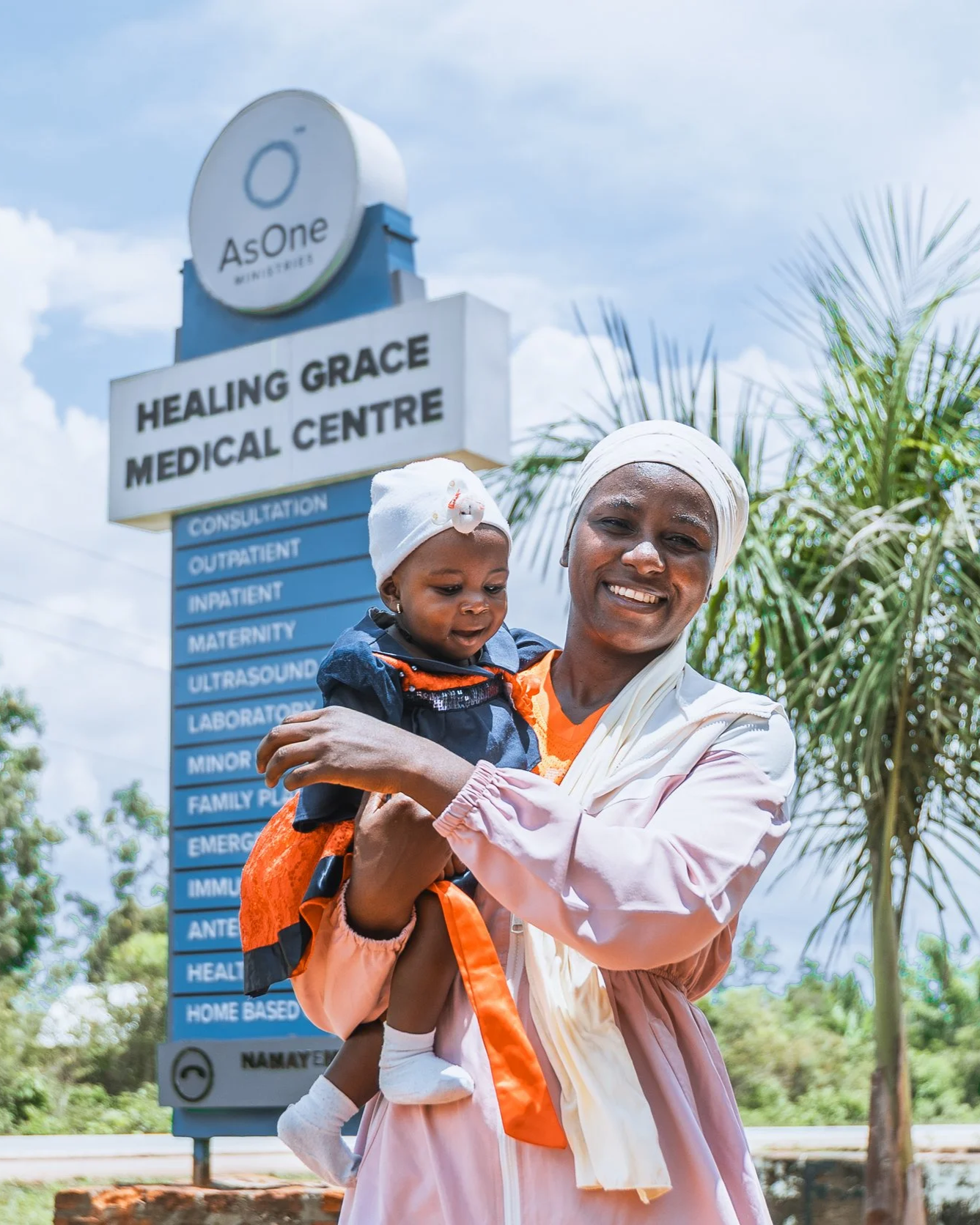 Mother holding her baby, Kateeba Azlin, an AsOne patient, in front of the Healing Grace Medical Centre sign in Namayemba