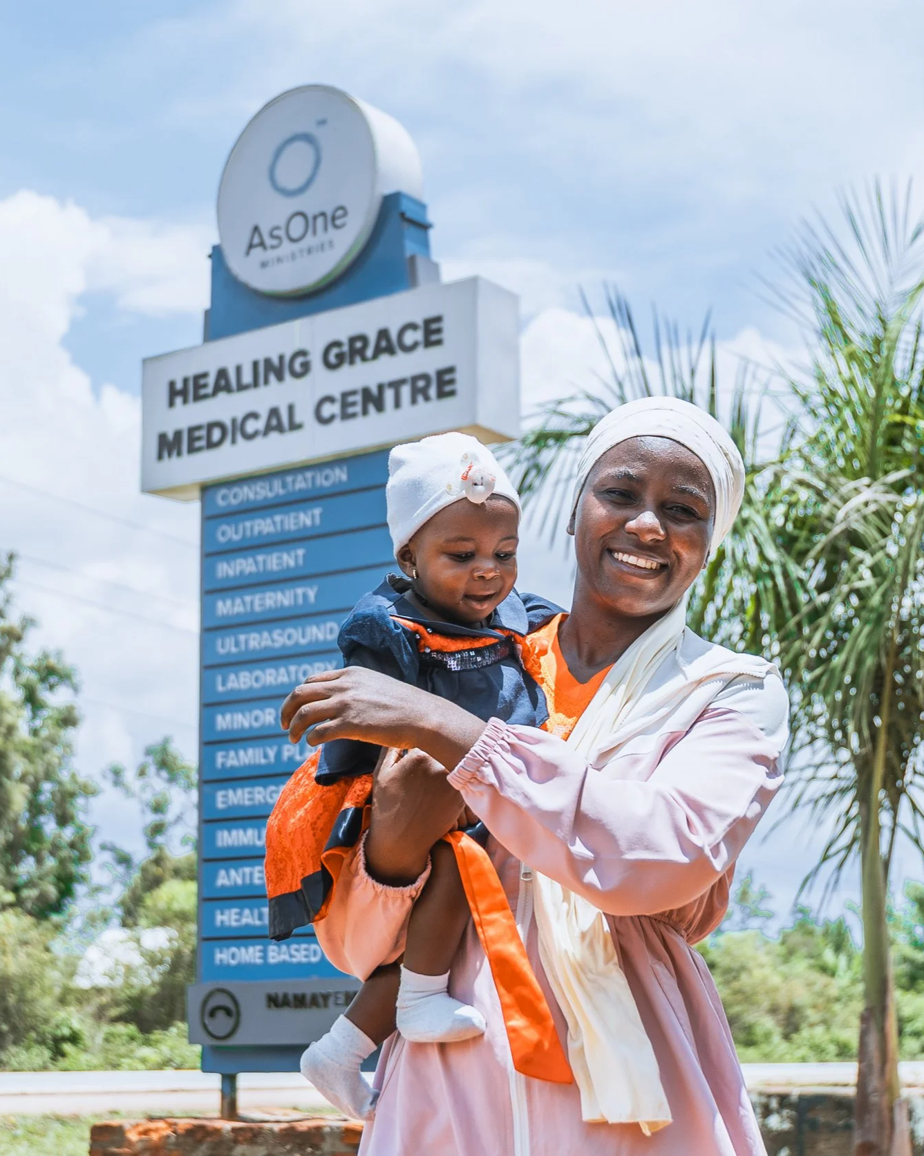 Mother holding her baby, Kateeba Azlin, an AsOne patient, in front of the Healing Grace Medical Centre sign in Namayemba