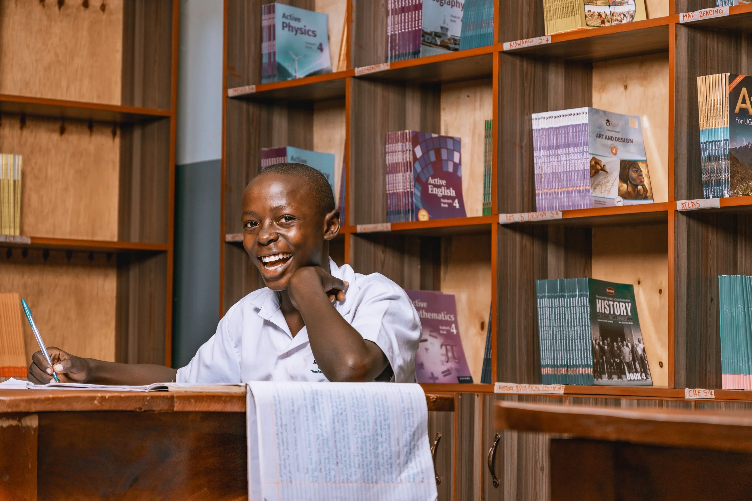 Smiling AsOne High School, Kaberamaido student writing at a desk in the school library