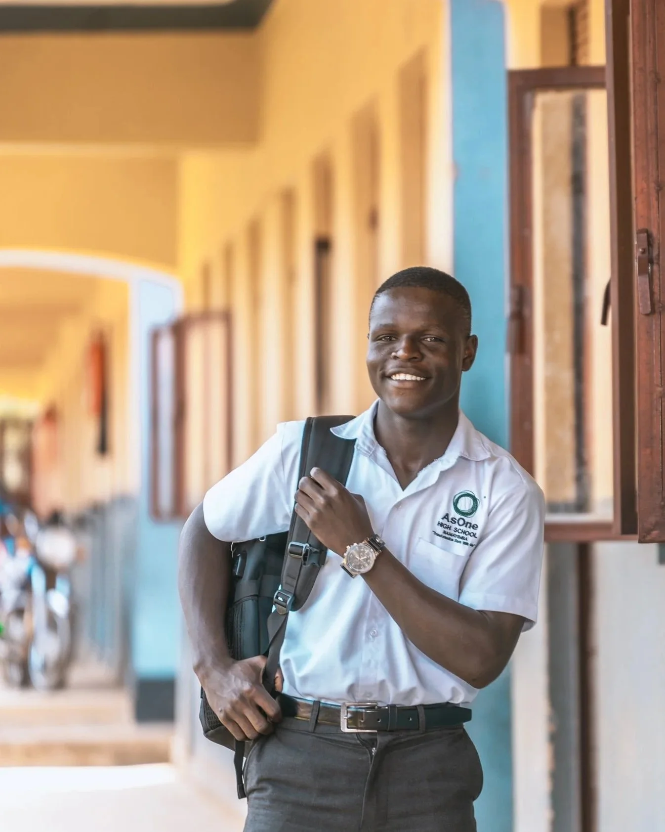 Smiling AsOne High School, Namayemba student in uniform carrying a backpack outside a school building