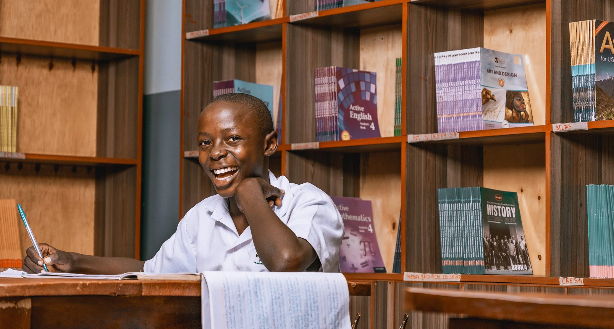 Smiling AsOne High School, Kaberamaido student writing at a desk in the school library