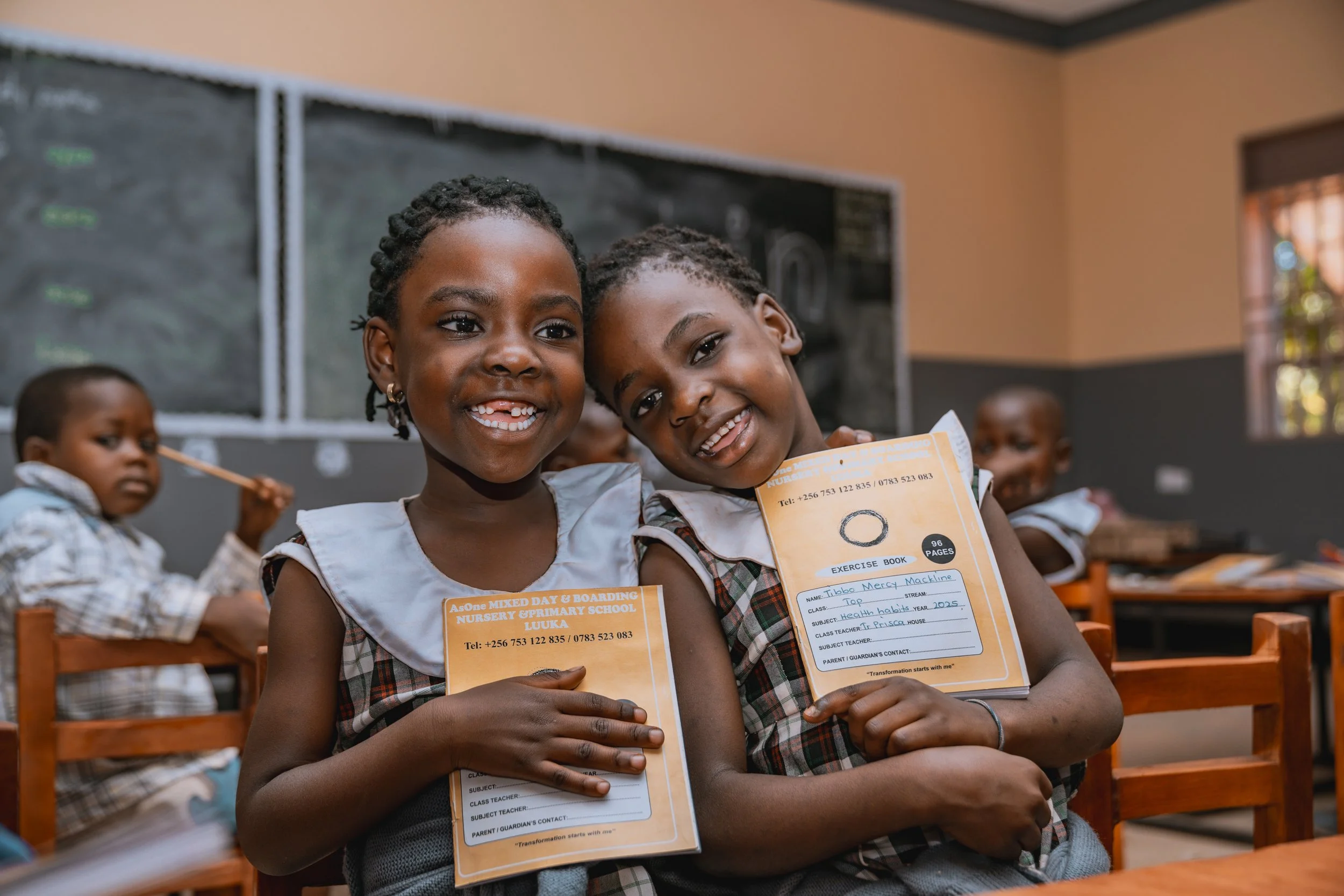Two smiling AsOne Nursery and Primary School, Luuka students in a classroom holding exercise books