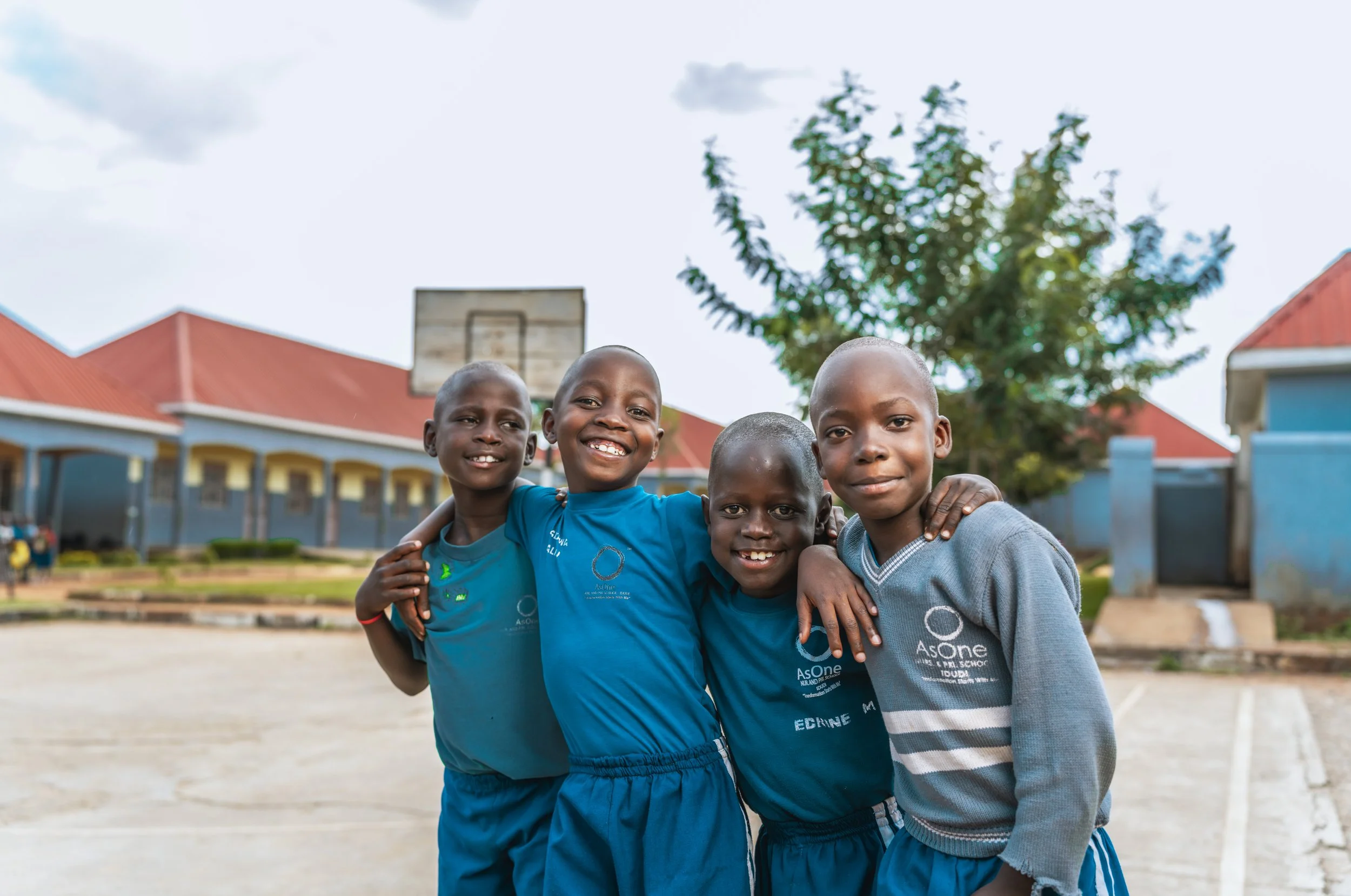 Four smiling pupils at AsOne Primary & Nursery School, Idudi, standing together in the school compound