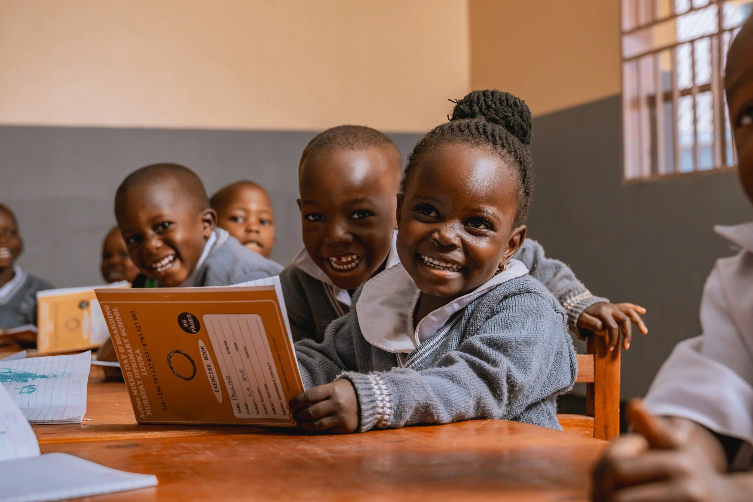 Smiling young students at desks in a classroom at AsOne Nursery & Primary School, Luuka