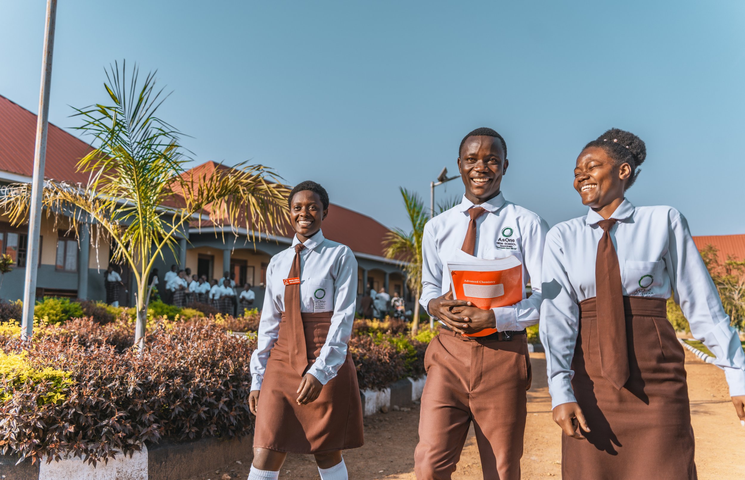 Three smiling AsOne High School, Namayemba students in uniform walking on campus, one holding a chemistry textbook