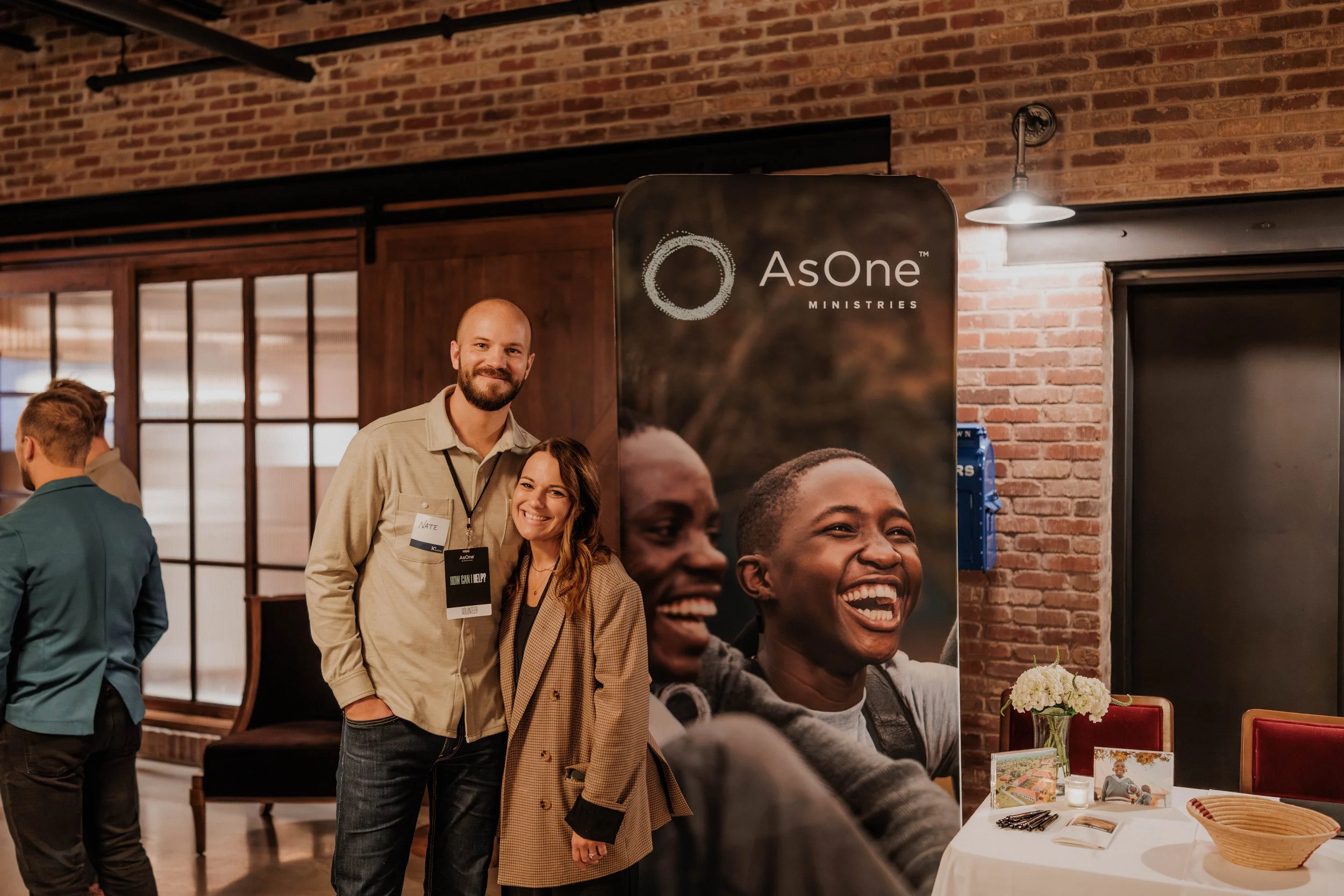 Two attendees smile for a photo beside an AsOne Ministries banner during the Denver event.
