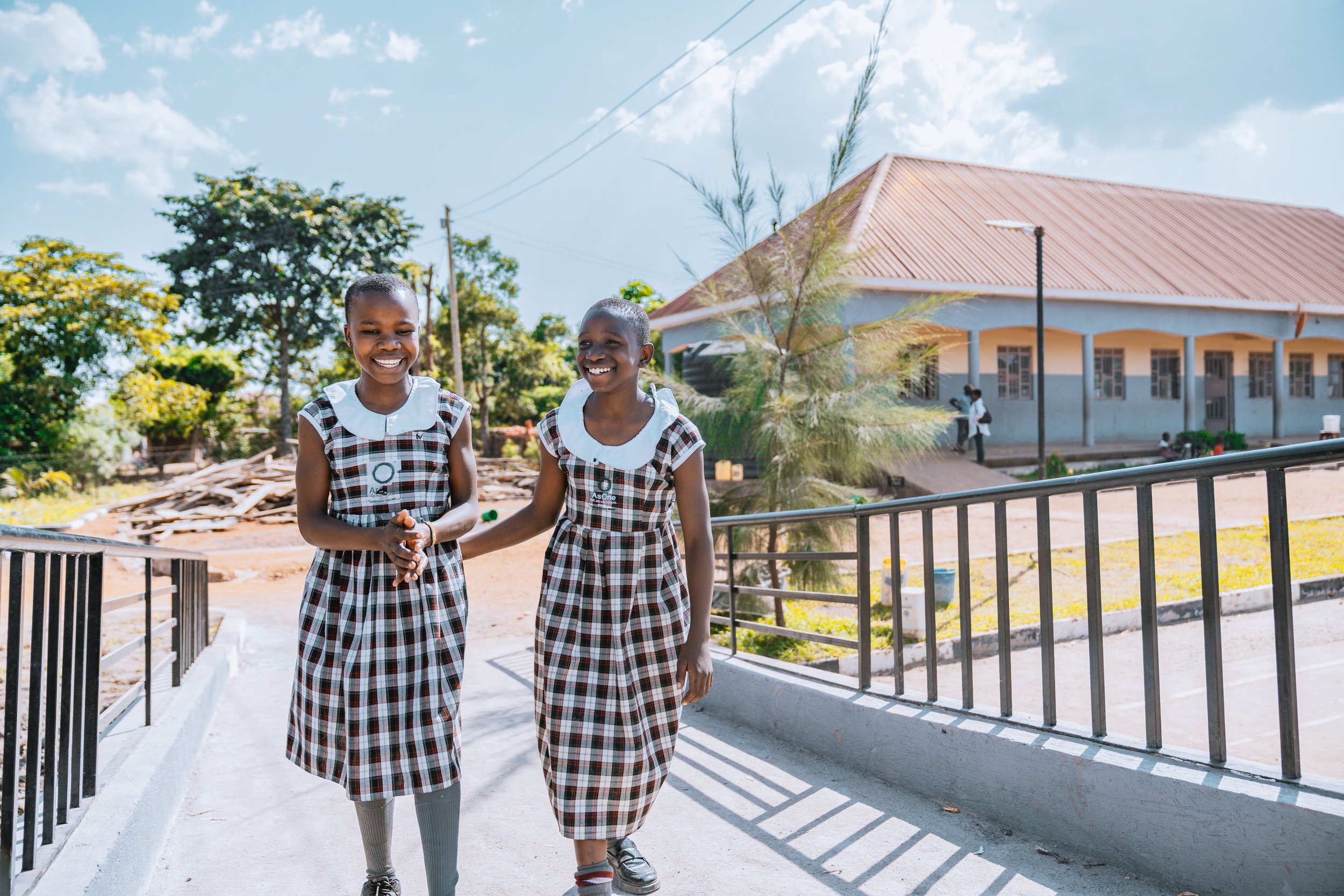 Two smiling AsOne Nursery & Primary School students in uniform posing in front of a campus building in Idudi