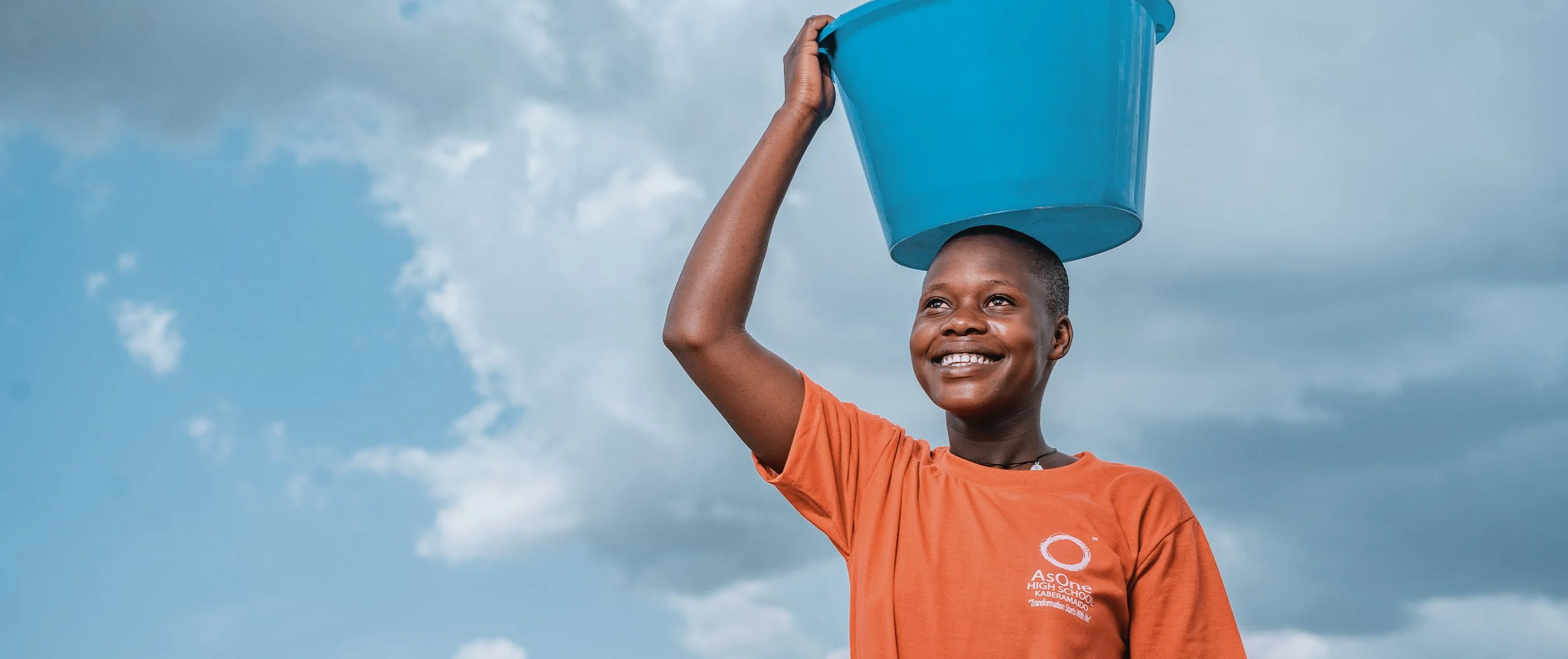 Smiling AsOne High School Kaberamaido student carrying a water bucket on her head.