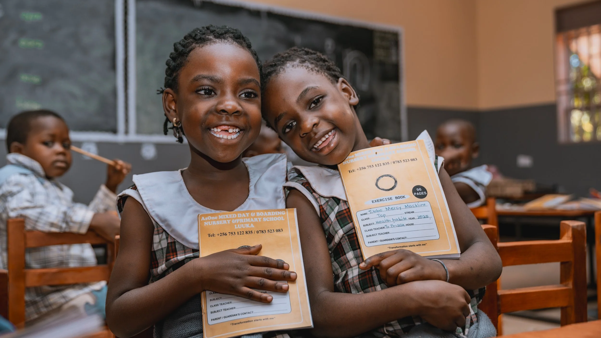 Two smiling AsOne Nursery and Primary School, Luuka students in a classroom holding exercise books
