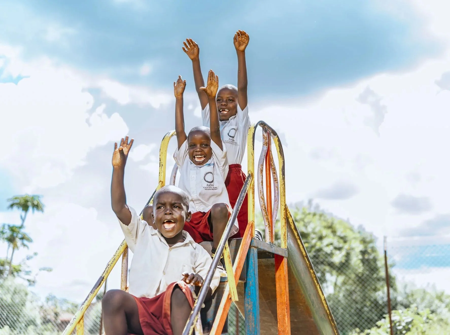 Three excited AsOne students from Kameramaido cheering with hands raised on a playground slide