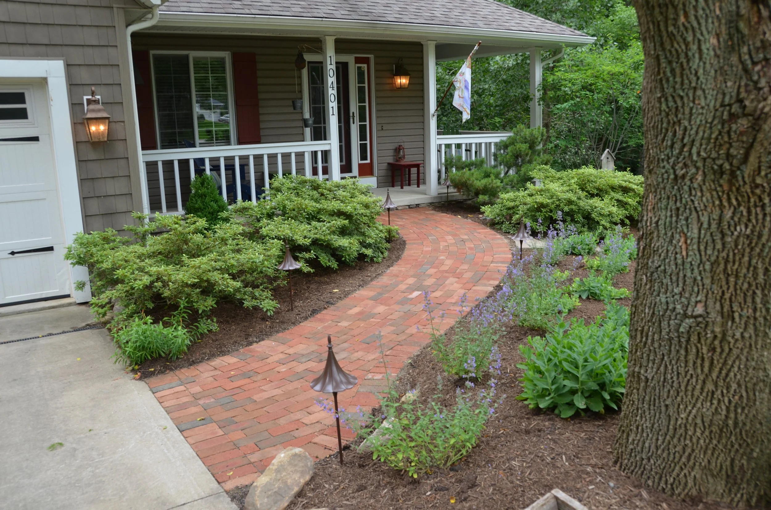 Front yard with a brick walkway, green shrubs, purple flowers, and a large tree. The house has a porch with a white railing, a red chair, and a flag hanging from a pole.