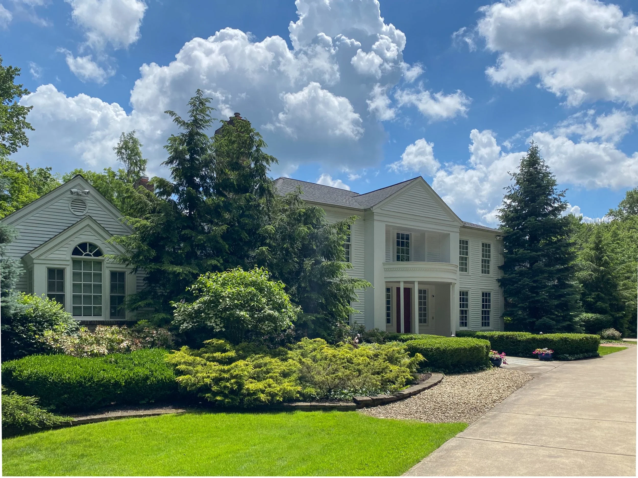 A two-story white house with large windows and a curved porch, surrounded by lush green trees and bushes under a partly cloudy sky, with a concrete driveway in the foreground.